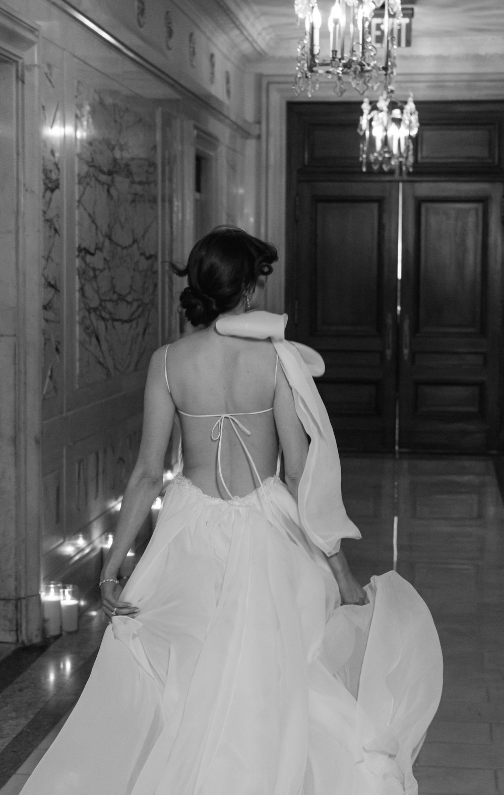 Elegant white wedding dress with open back and flowing train captured in luxurious hallway with chandeliers.