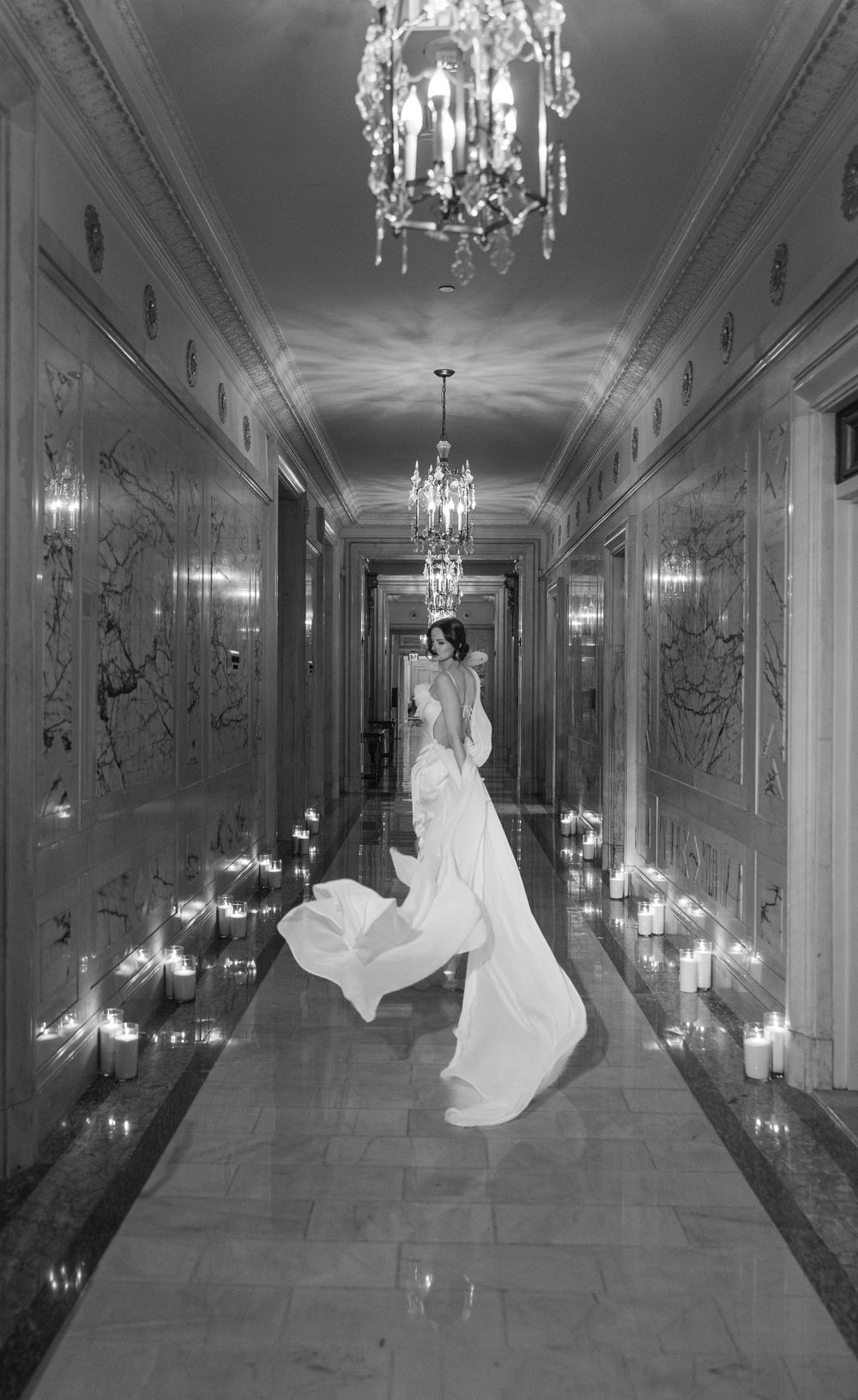Black and white photograph of flowing wedding gown in chandelier-lit corridor.