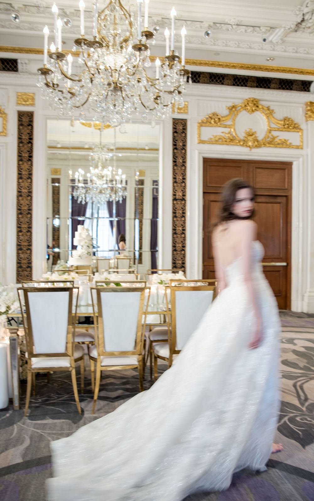 Motion-blurred wedding dress photo in ornate room with gold details and crystal chandelier.