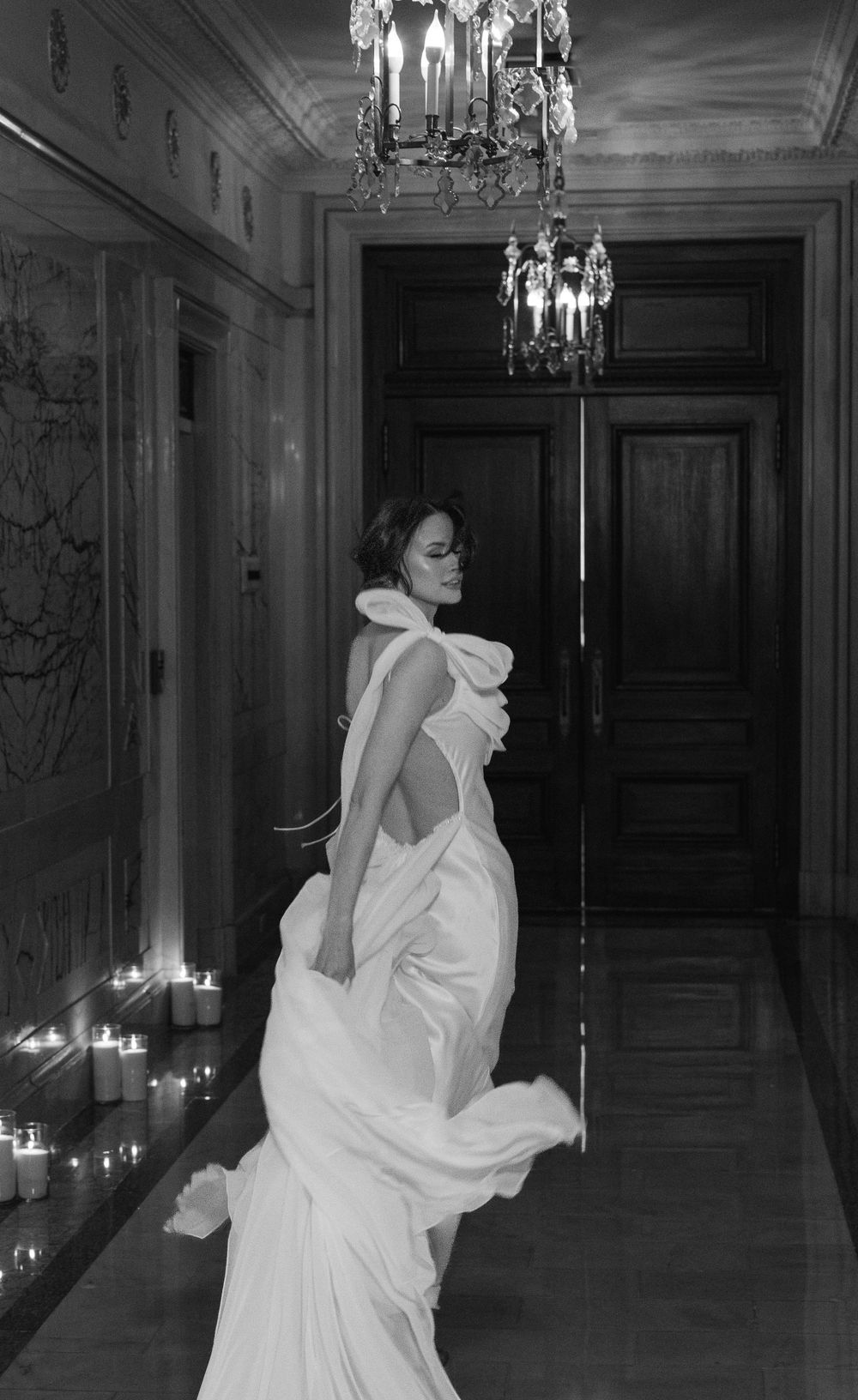 Black and white photo of flowing backless wedding dress in candlelit hallway with crystal chandeliers.
