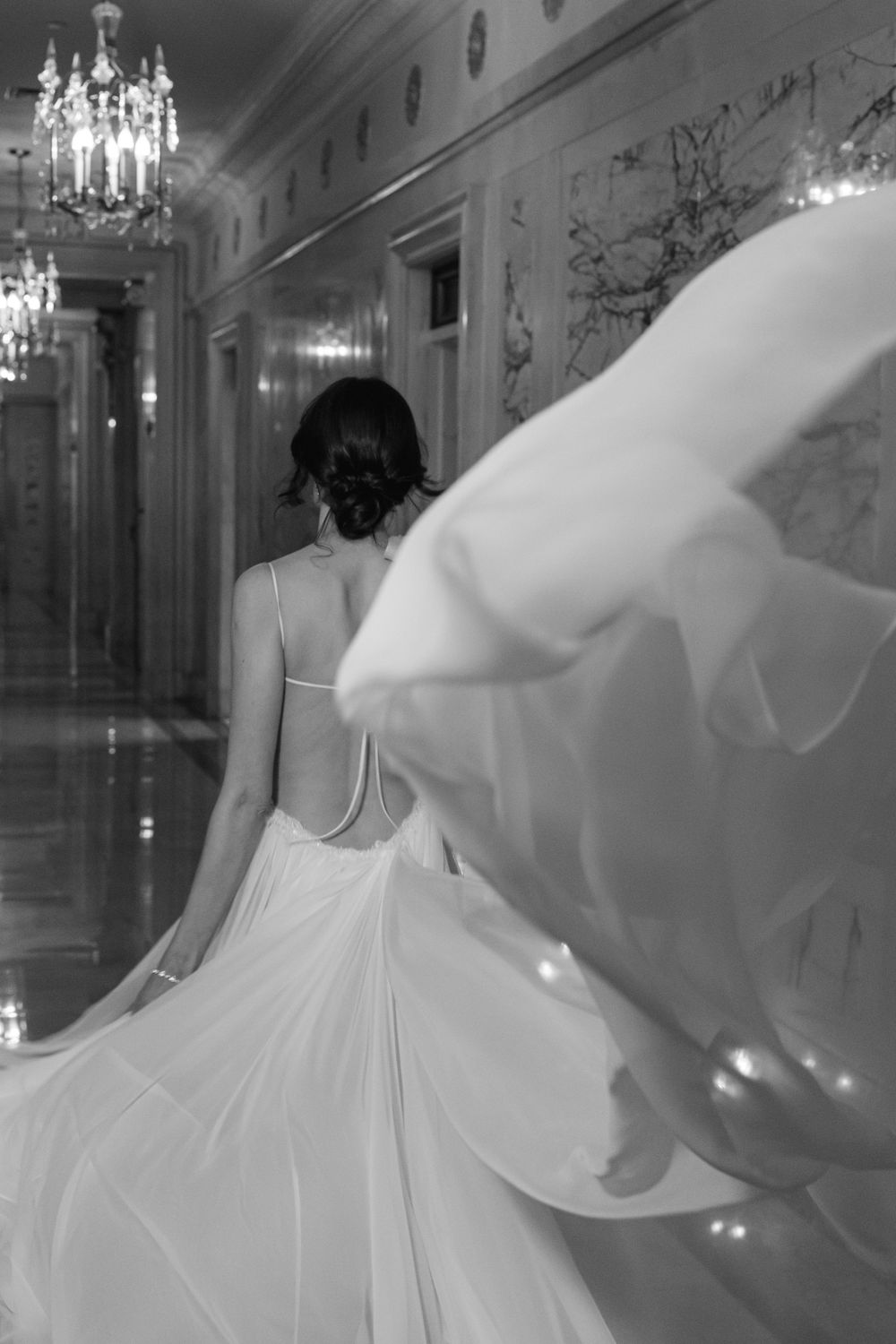 Back view of wedding dress flowing dramatically in marble hallway with crystal chandeliers.