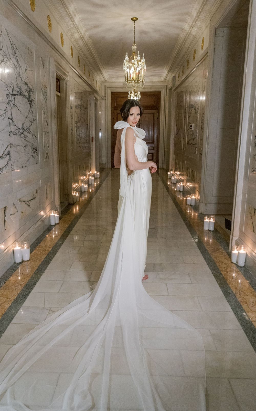 Elegant white wedding gown with flowing train shown in marble hallway lined with glowing candles and crystal chandelier.