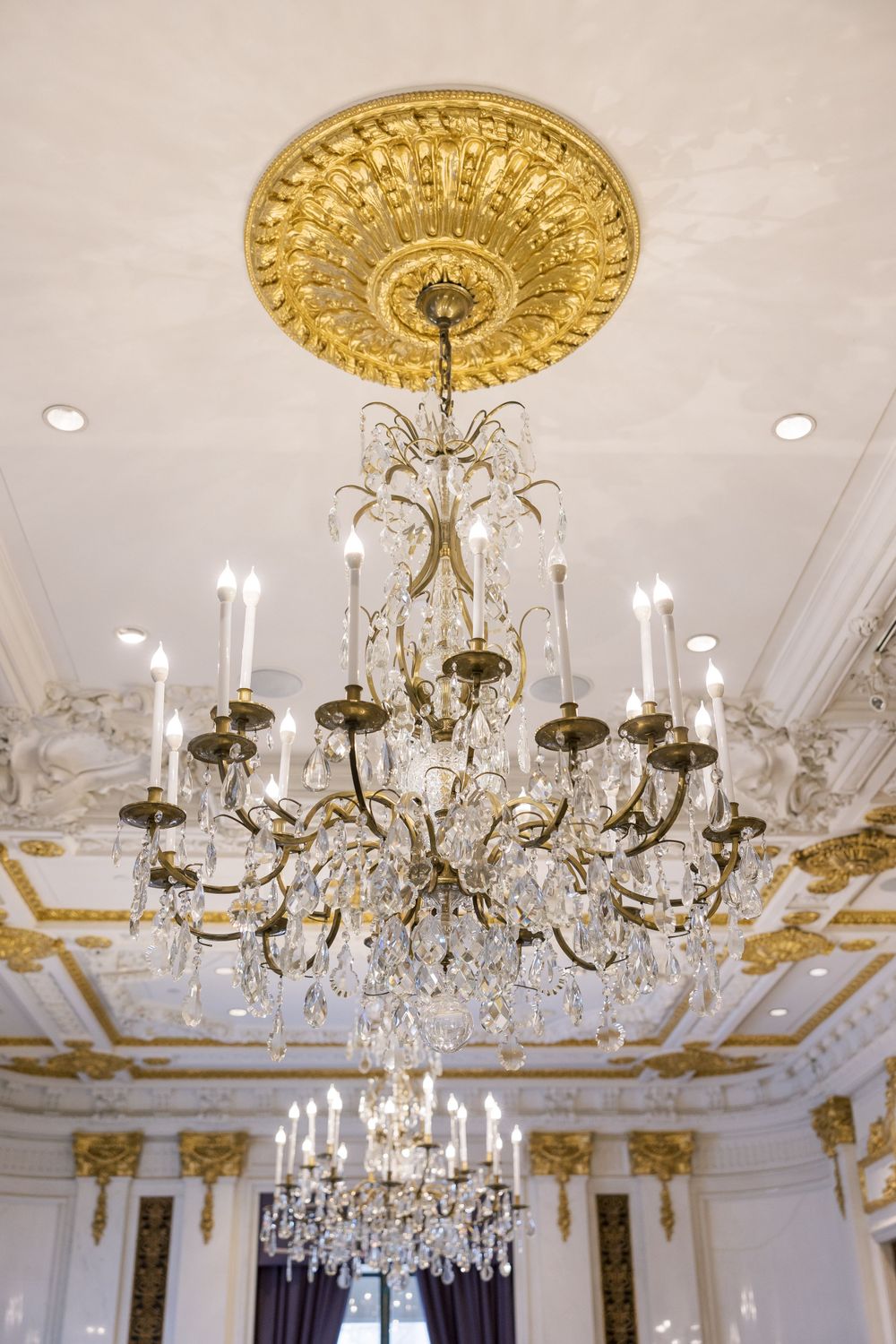 Crystal chandelier hanging from gold ceiling medallion in an elegant white and gold room.