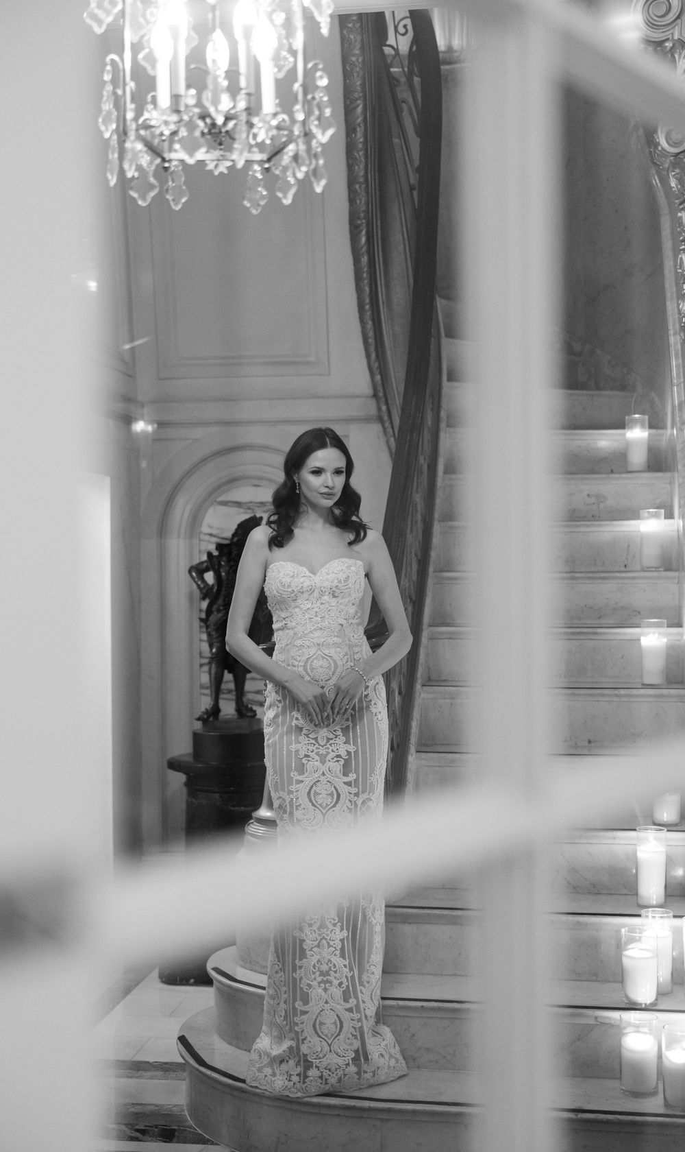 A bride wearing an elegant lace gown poses in a luxurious interior with a chandelier and ornate details.