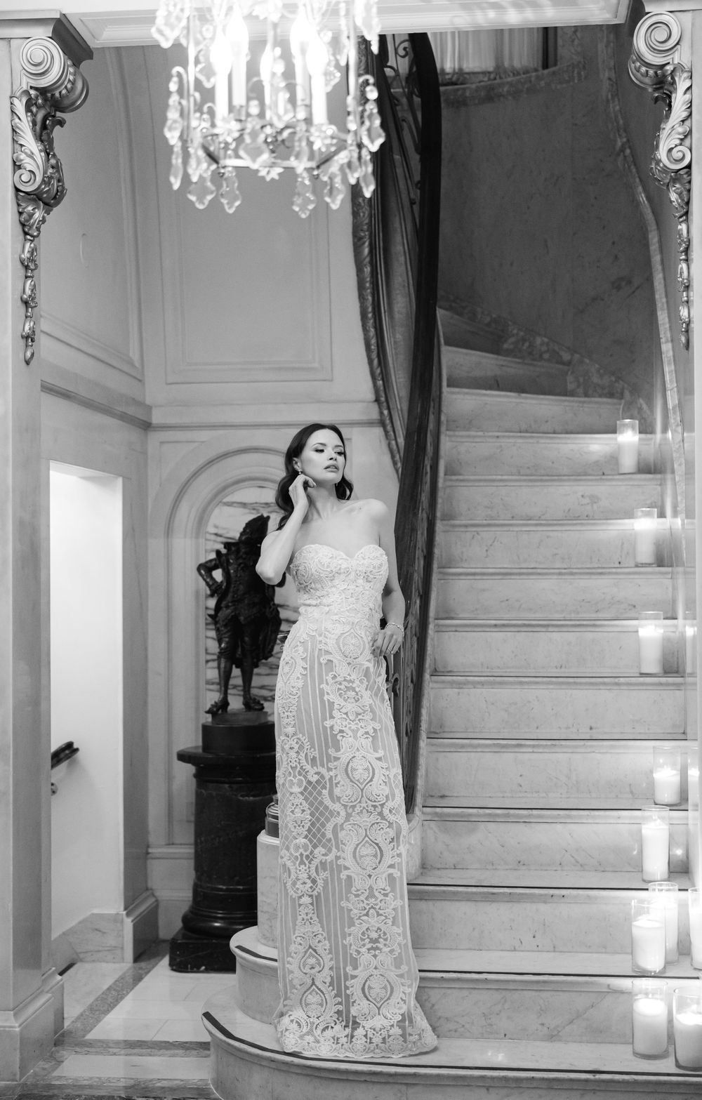 Black and white photo of bride in lace gown posing on grand staircase with chandelier.