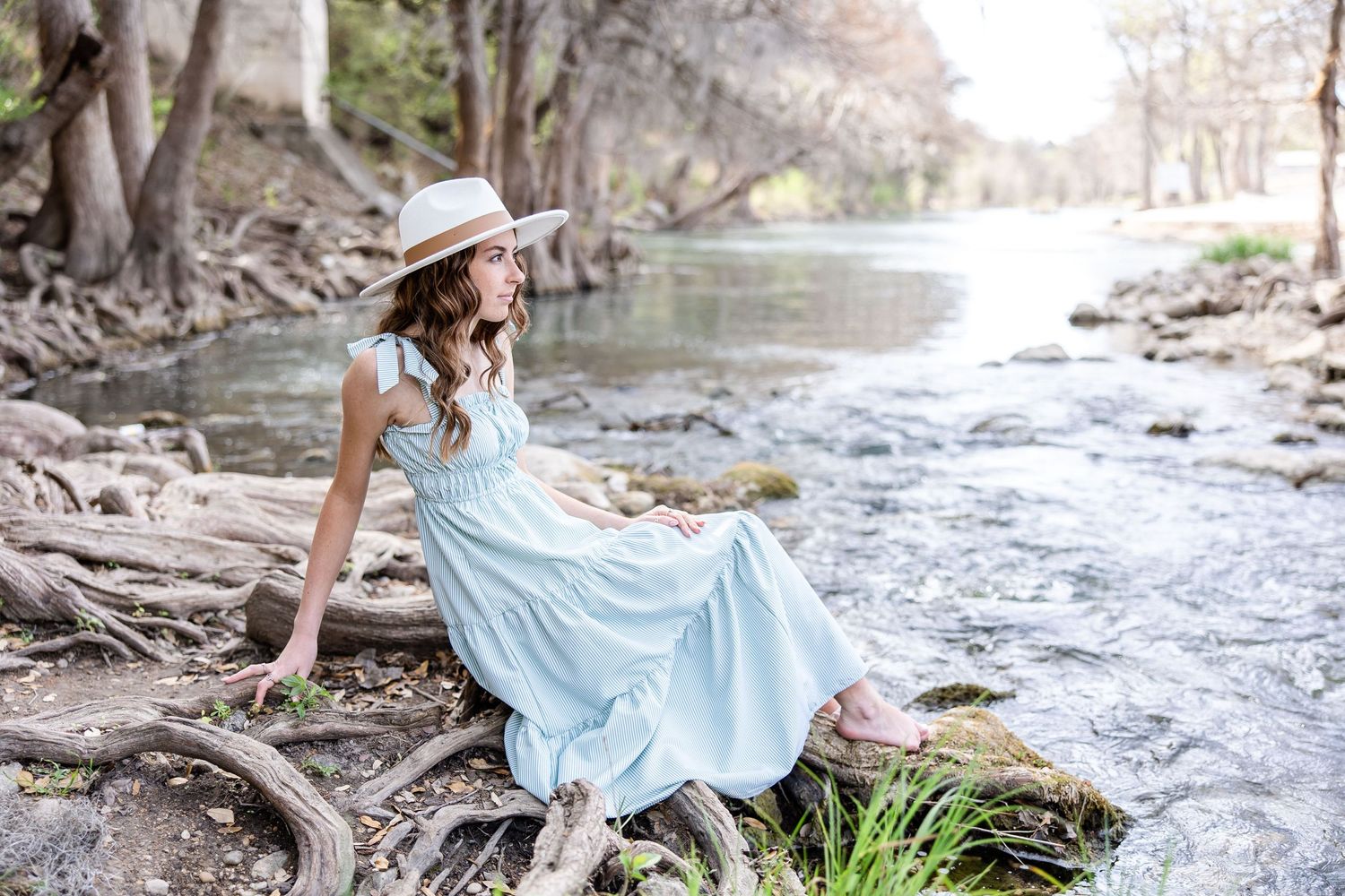 Woman in flowing blue dress sits by a peaceful stream wearing a sunhat in springtime.