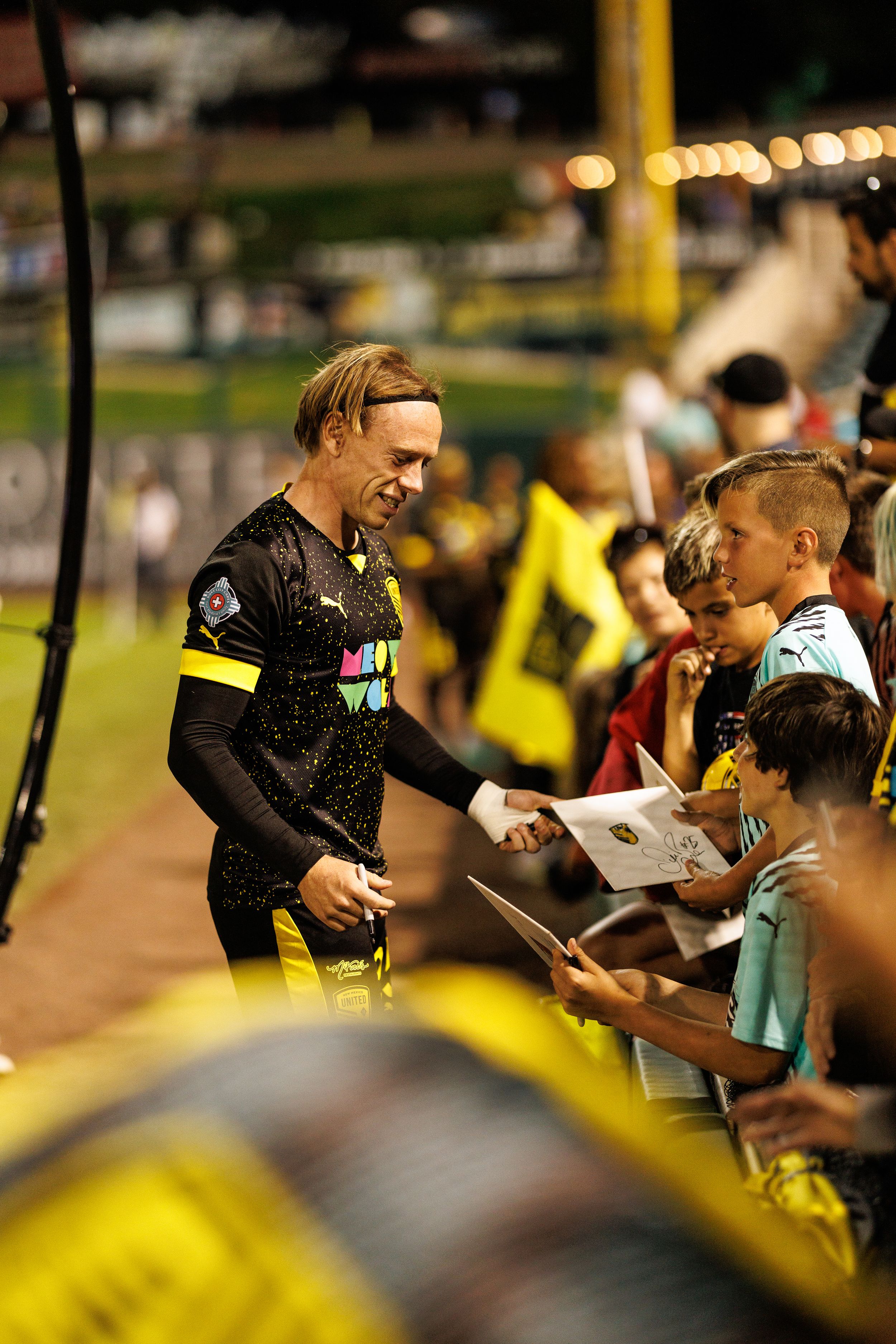 Player signs autographs for fans along stadium sideline during evening game.
