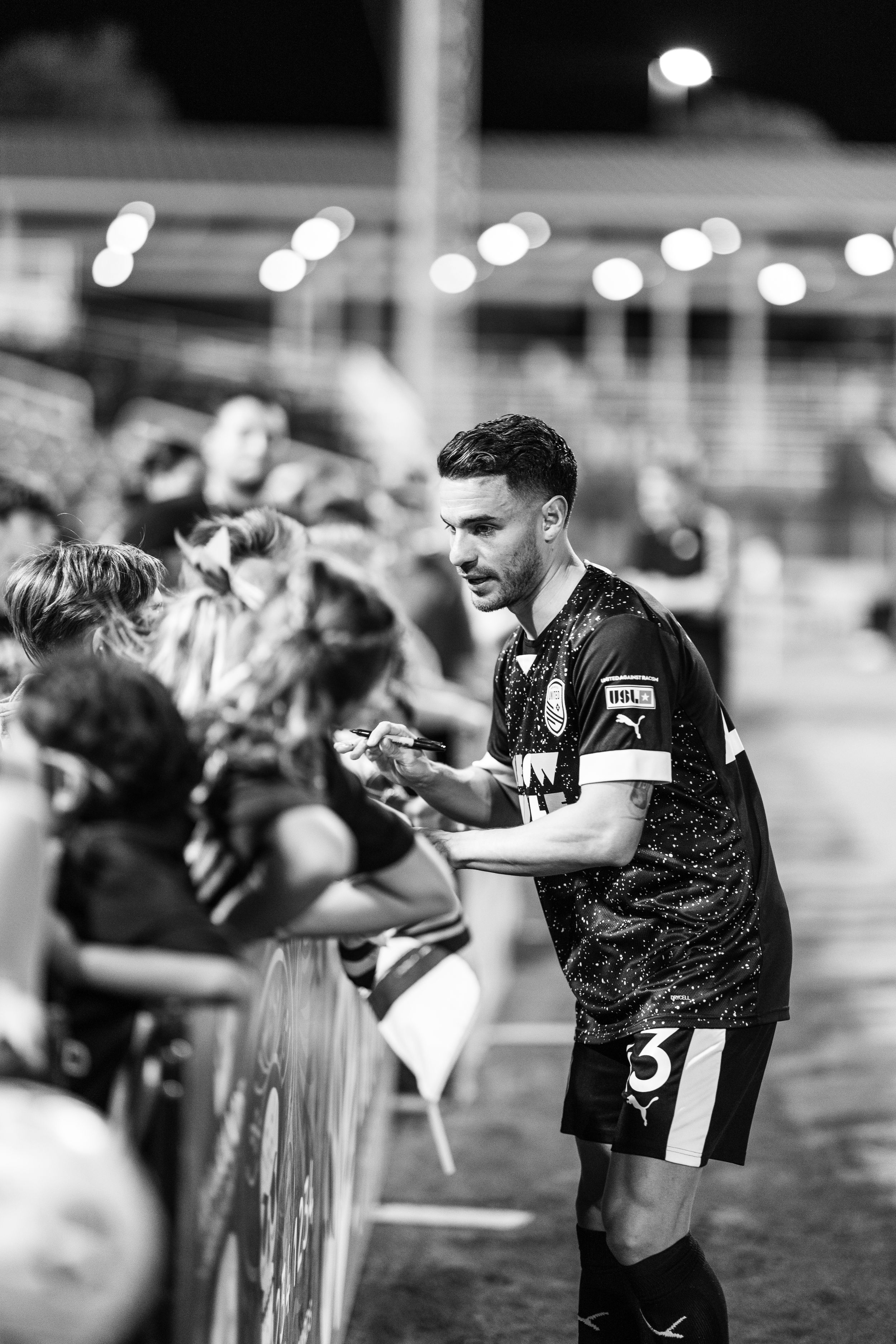 Player in dark uniform signs autographs for waiting fans along stadium barrier.