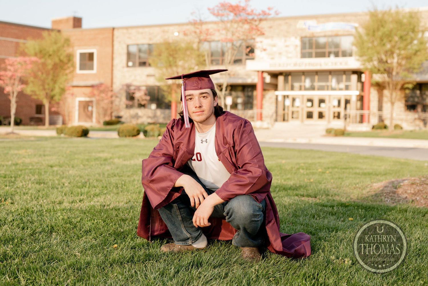 Graduate poses in burgundy cap and gown on campus lawn with academic building in background.
