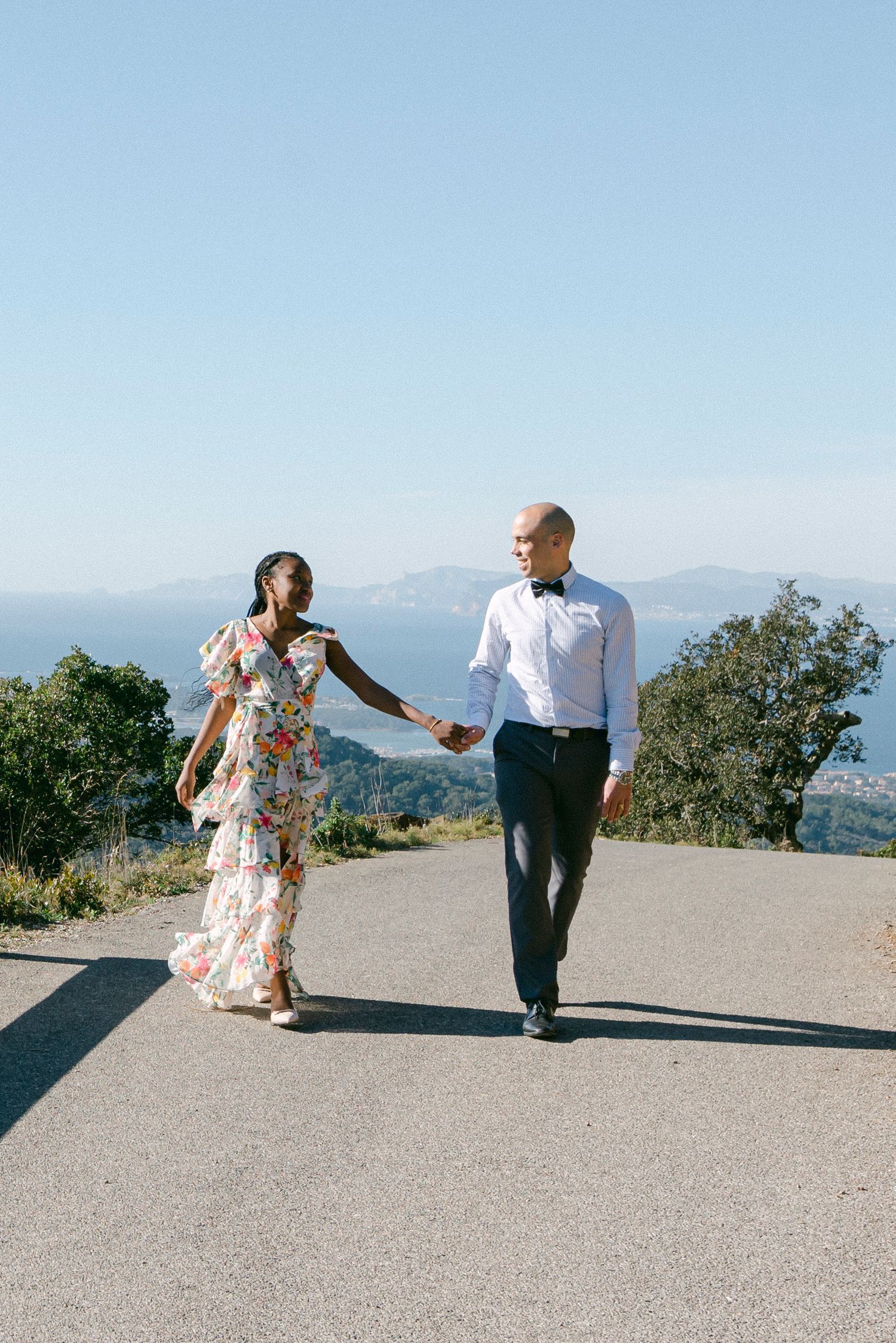 A couple strolls together on a mountaintop path overlooking the ocean and distant mountains.