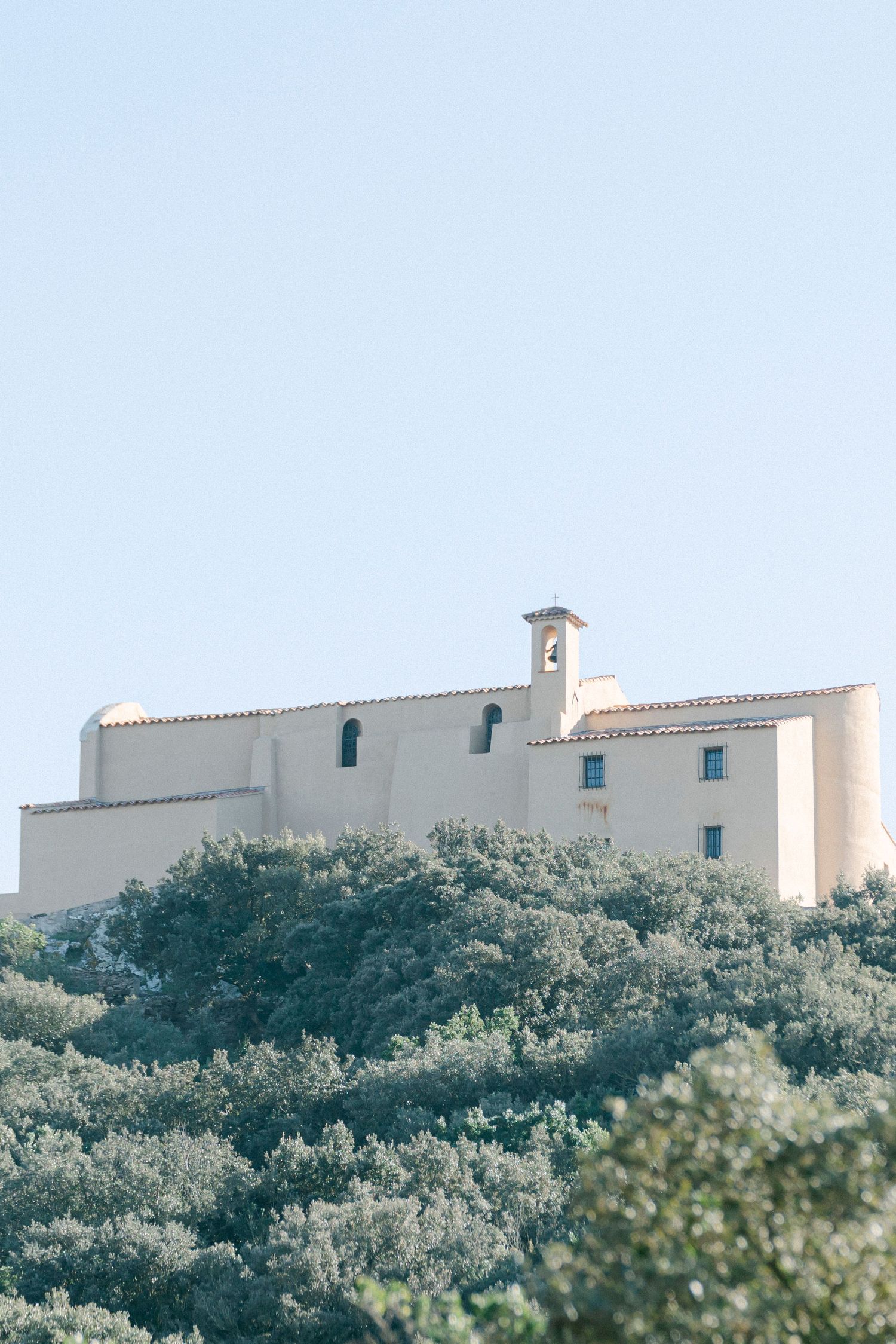 White stone monastery or church perched on a hilltop surrounded by lush Mediterranean vegetation.
