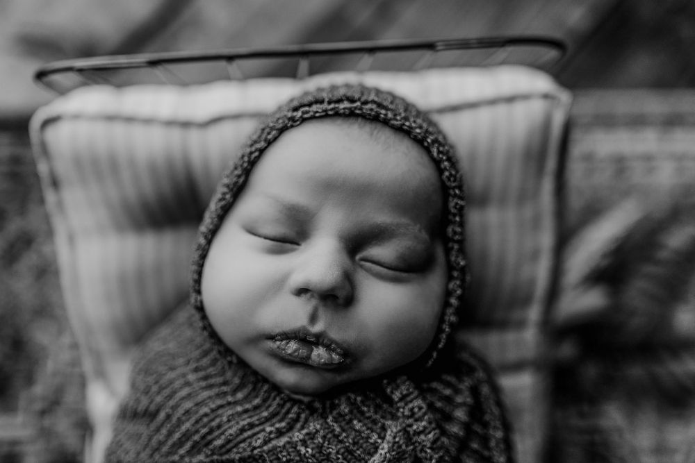 A black and white portrait of a sleeping baby wearing a knitted bonnet and sweater.