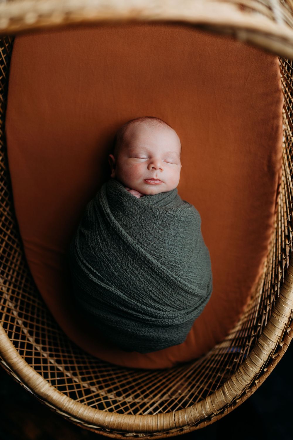 A sleeping newborn swaddled in a dark green wrap resting in a woven rattan basket.