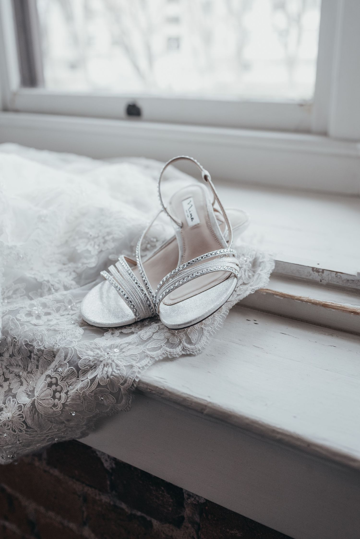 White bridal sandals with rhinestone details rest on a windowsill next to lace fabric.