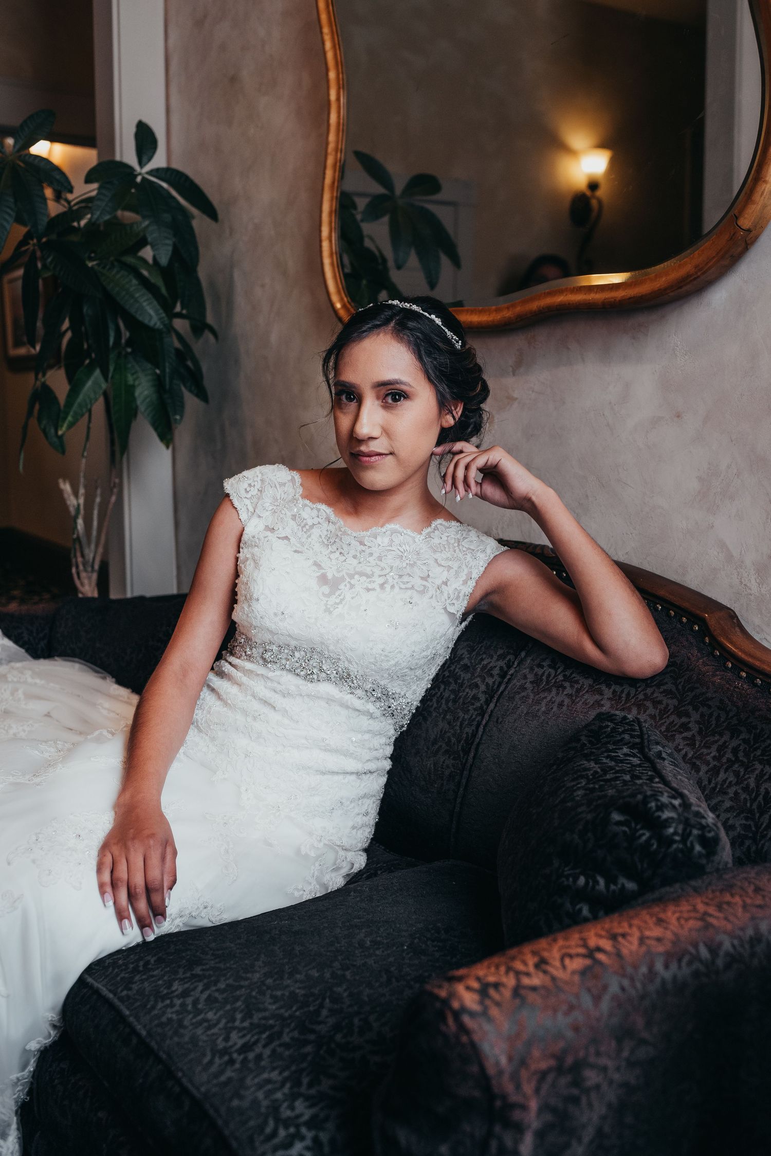 Elegant bridal portrait featuring a white lace wedding dress against a dark vintage couch with houseplants in background.