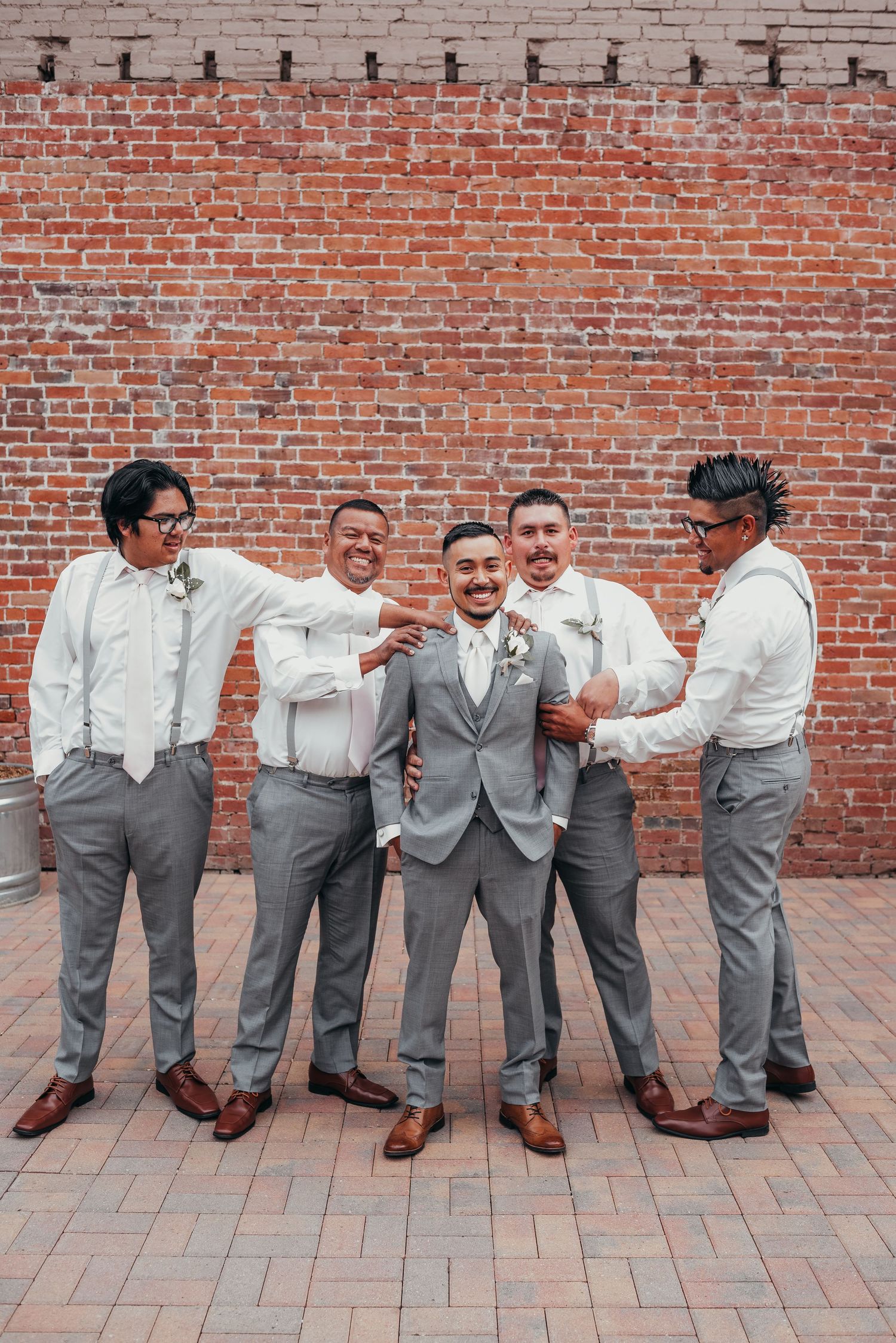 Groomsmen in grey suits and suspenders pose playfully against a brick wall.