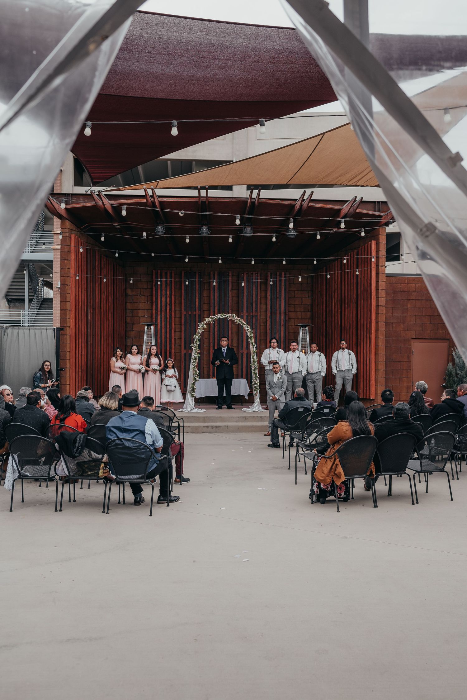 An outdoor wedding ceremony setup with wooden backdrop, string lights, and rows of chairs for guests.