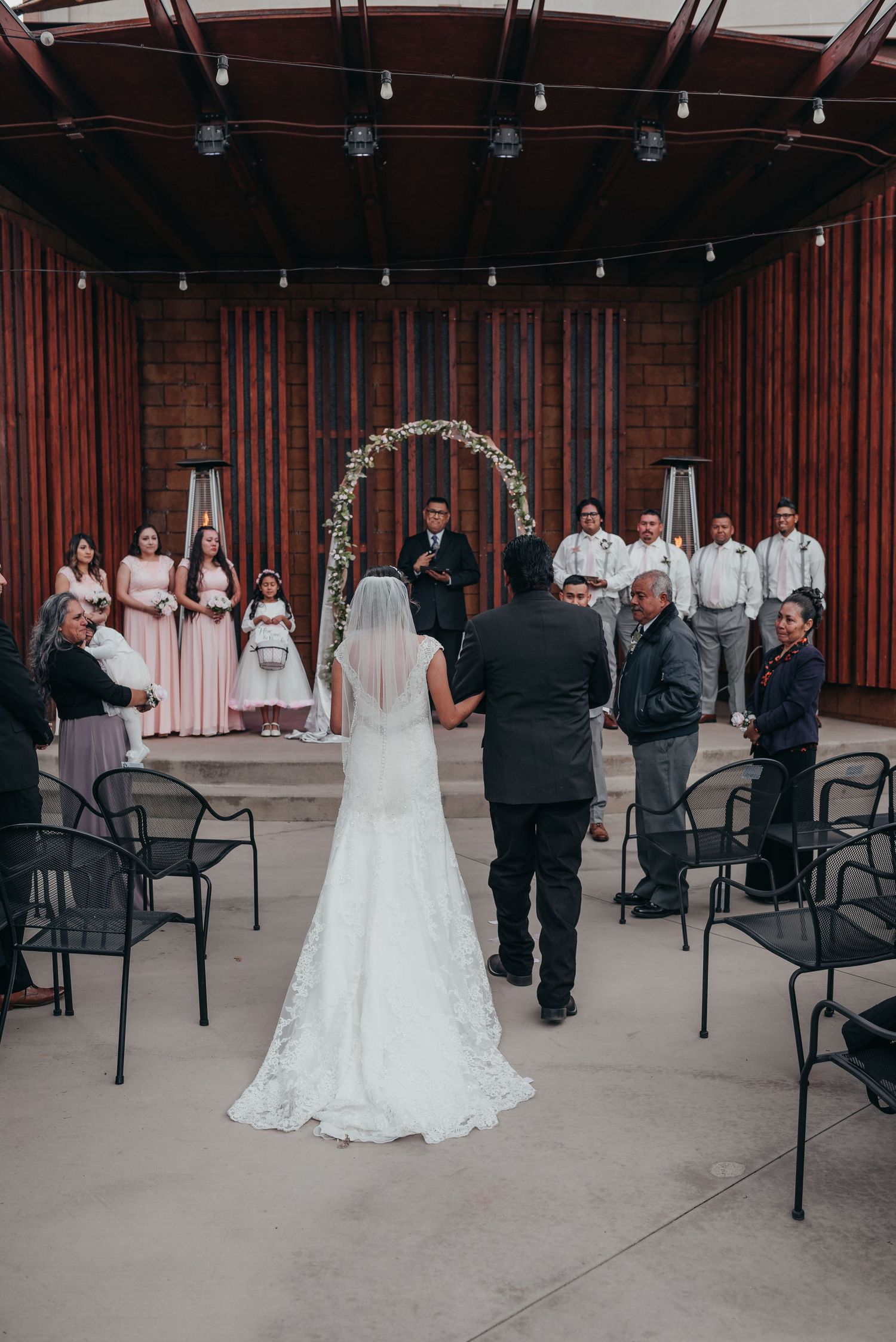A wedding ceremony in progress at an outdoor venue with wooden panels and floral arch.