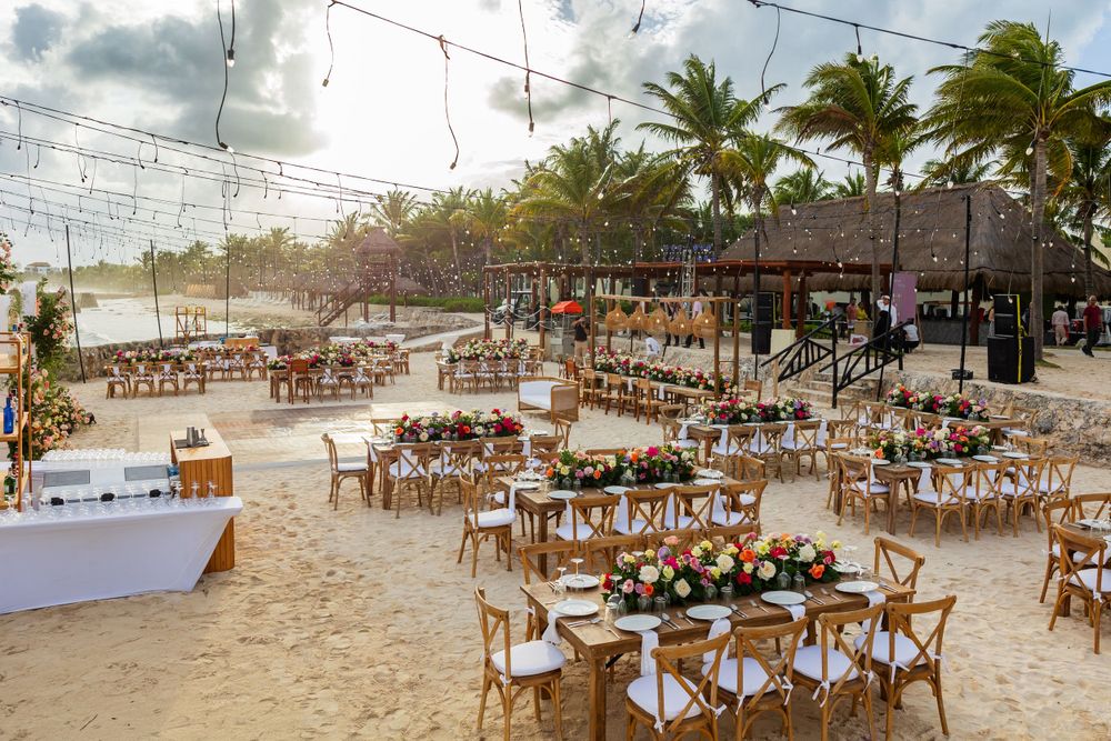 Beachfront wedding reception setup with wooden chairs and string lights under palm trees.