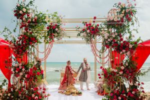 Ornate wedding ceremony arch with red roses and florals on a beach with the ocean in the background.