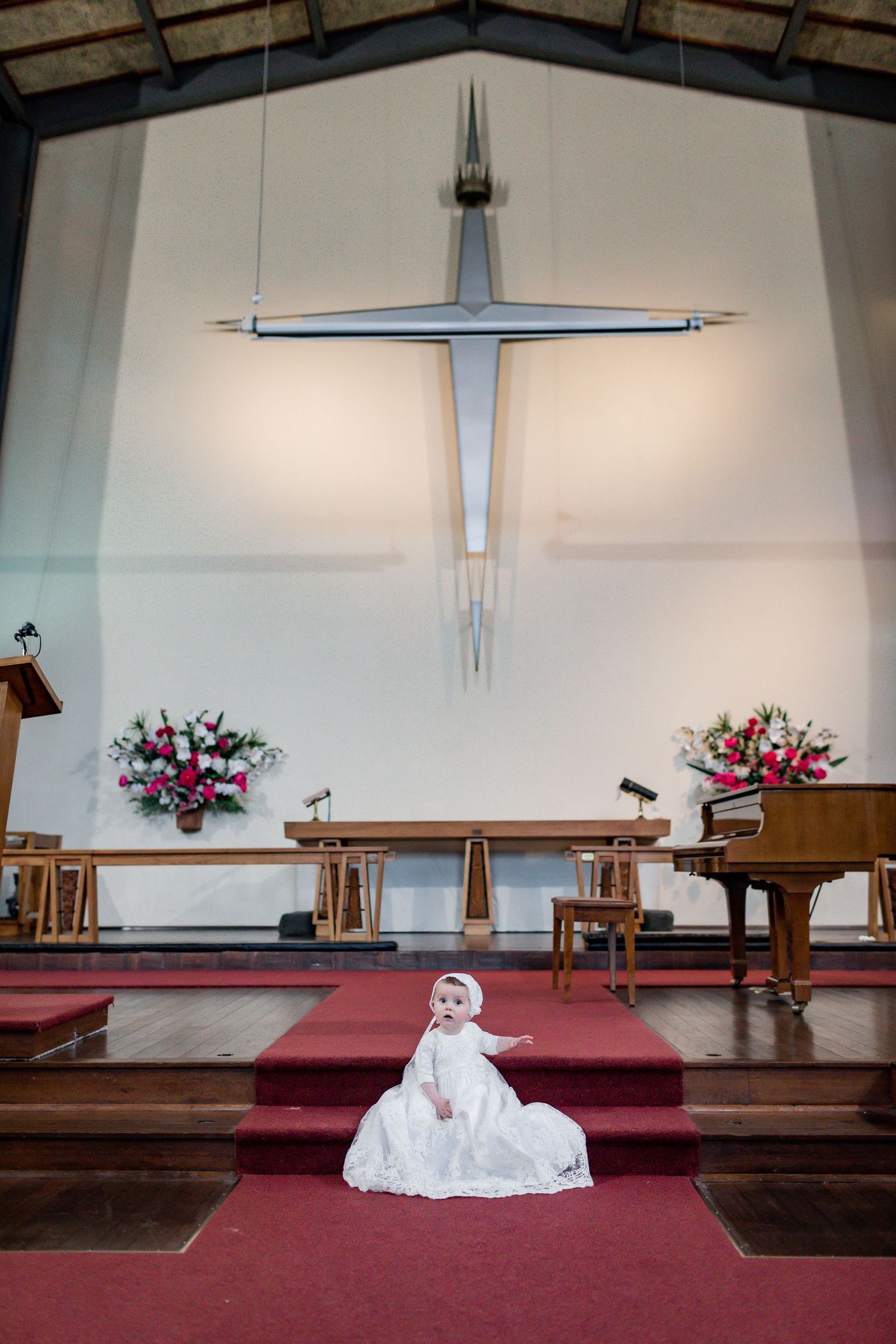 White christening gown displayed on red carpeted church altar under large modern cross and floral arrangements.