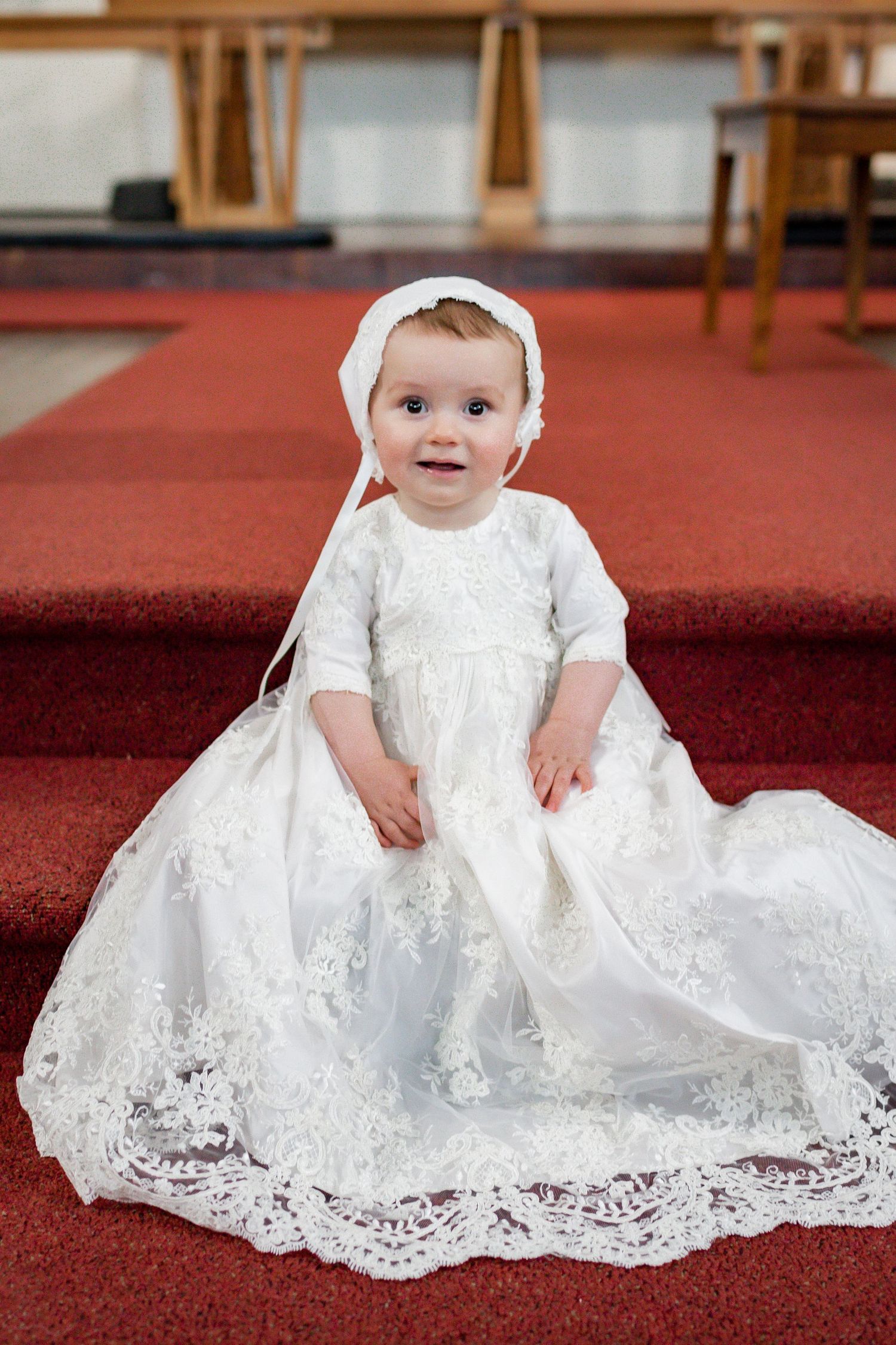 Baby wearing traditional white christening gown and bonnet sits on red church carpet.