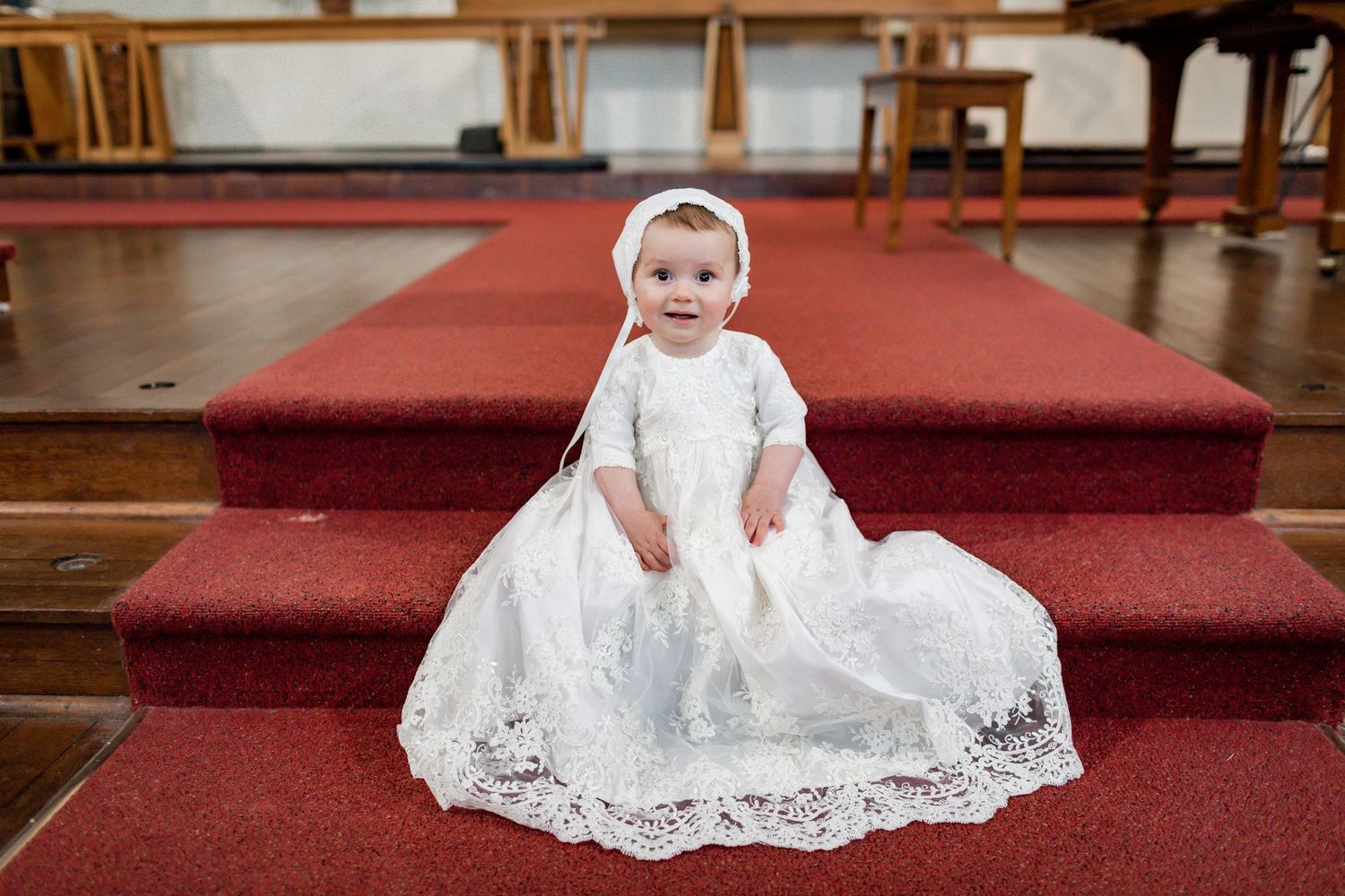 Baby in ornate white lace christening gown sits on red carpeted church steps.
