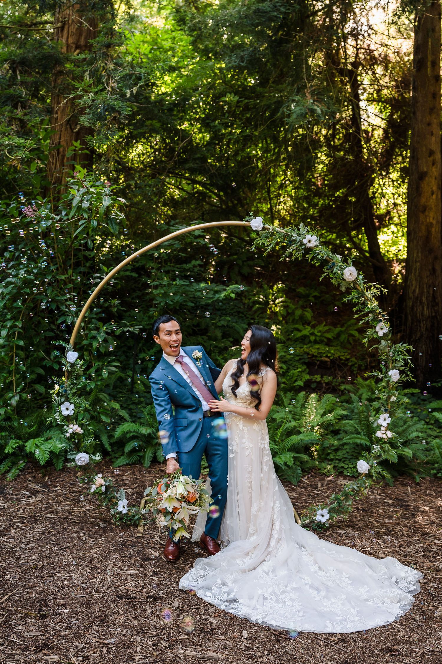 A couple stands under an elegant floral arch in a wooded setting with trailing white dress.