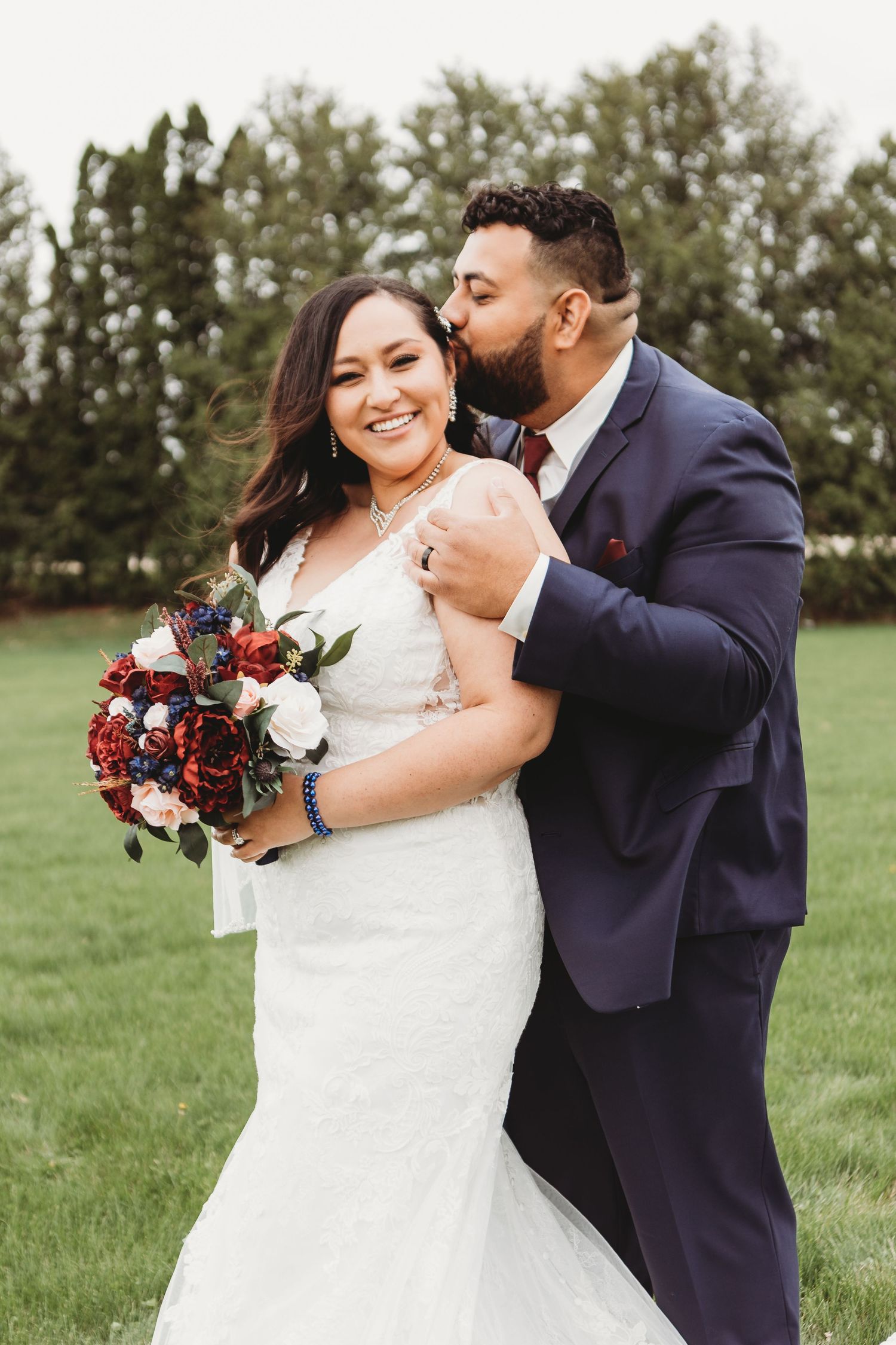 Wedding couple embracing outdoors with bride holding a burgundy and navy floral bouquet.