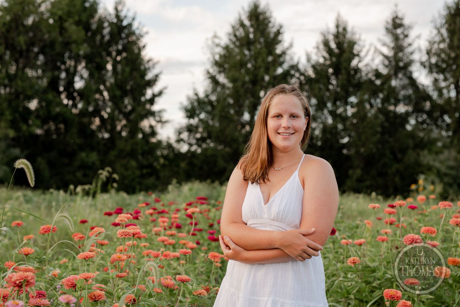A woman in a white sundress poses confidently in a field of red poppies with evergreen trees in the background.