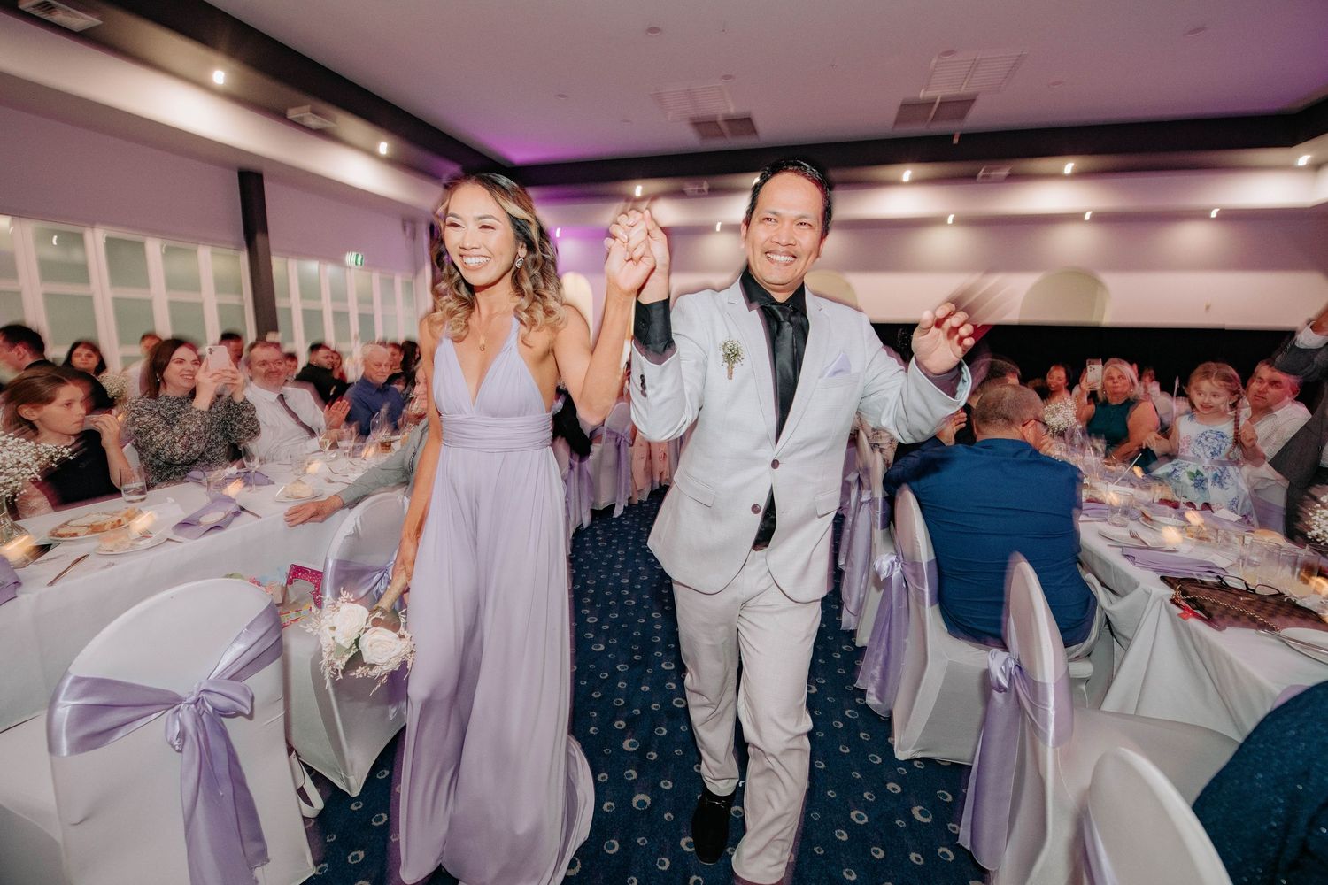 Wedding party members dance joyfully in a reception hall with decorated tables and guests.