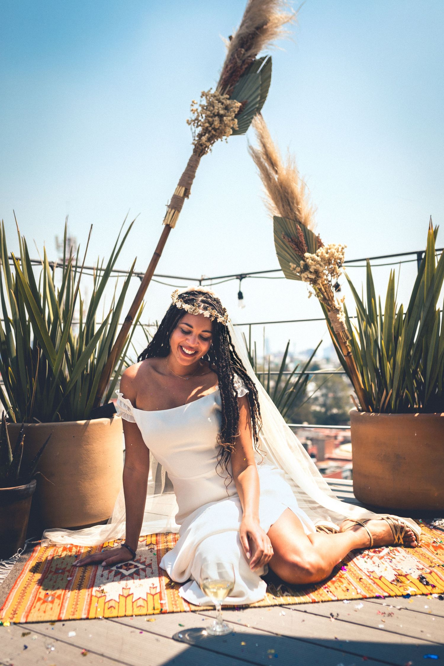 Person in white dress relaxing on orange blanket with potted plants and champagne.
