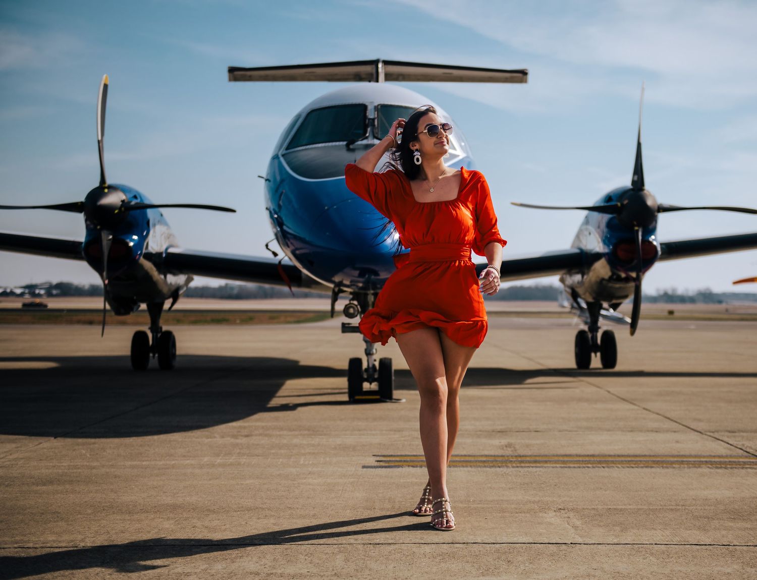 Red dress flowing in breeze between parked aircraft on sunny airport tarmac.