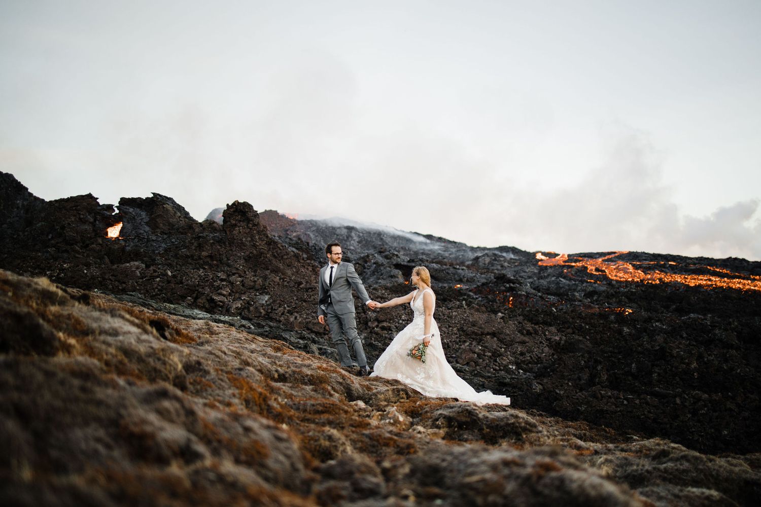 A couple in wedding attire poses dramatically on volcanic rocks at sunset.