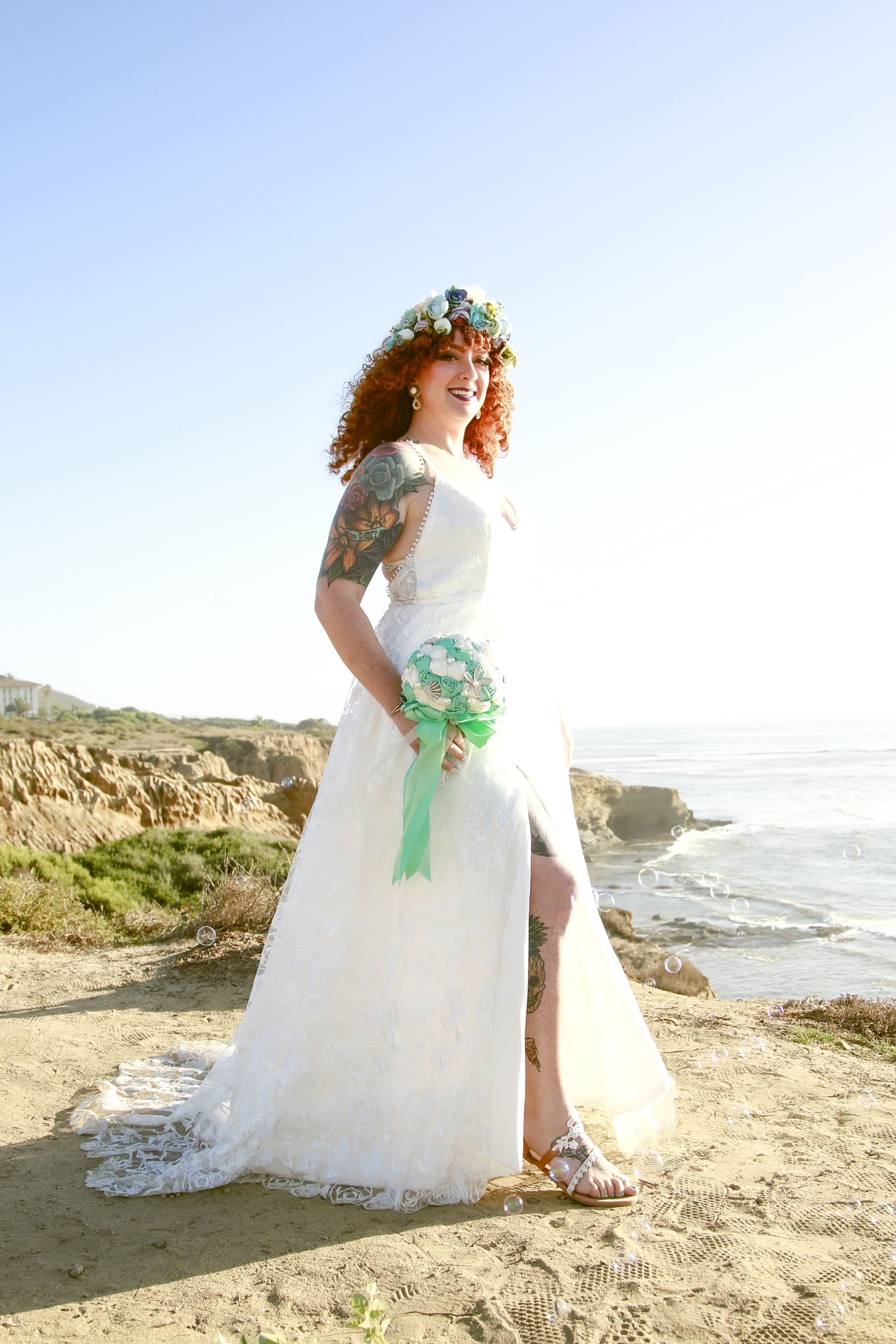 A bride in a flowing white dress poses on a beach with ocean waves in the background.