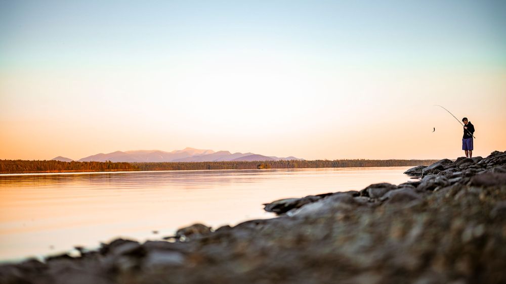 Lone figure fishing on a rocky shoreline during golden hour with mountains in the distance.