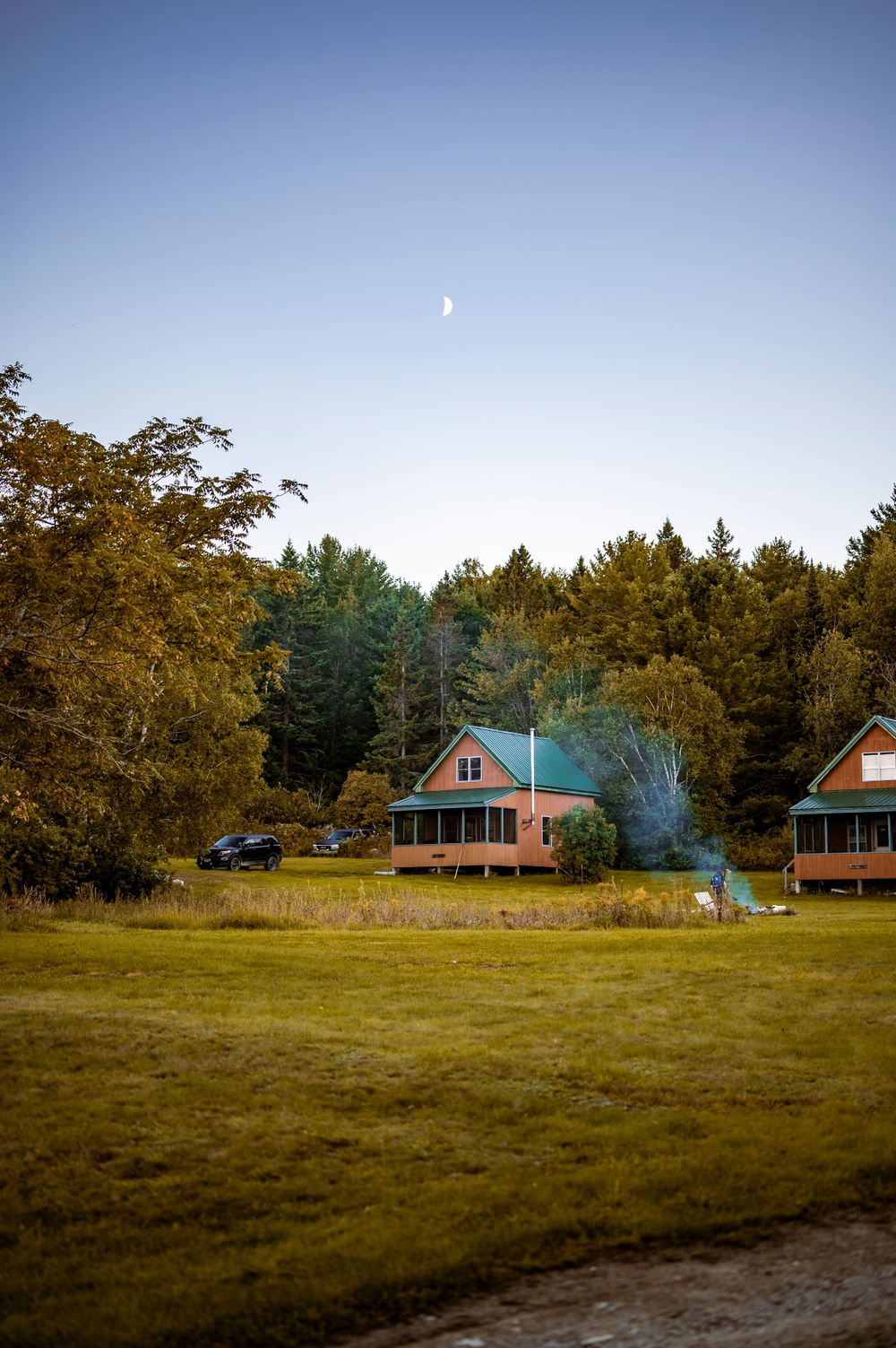 Rustic cabin nestled in autumn woods with a crescent moon visible in the blue sky.