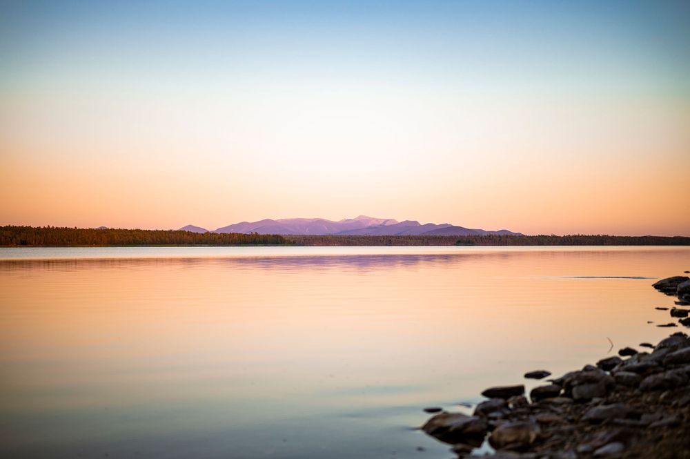 Peaceful sunset reflection on a still lake with rocky shore and purple mountains in the distance.