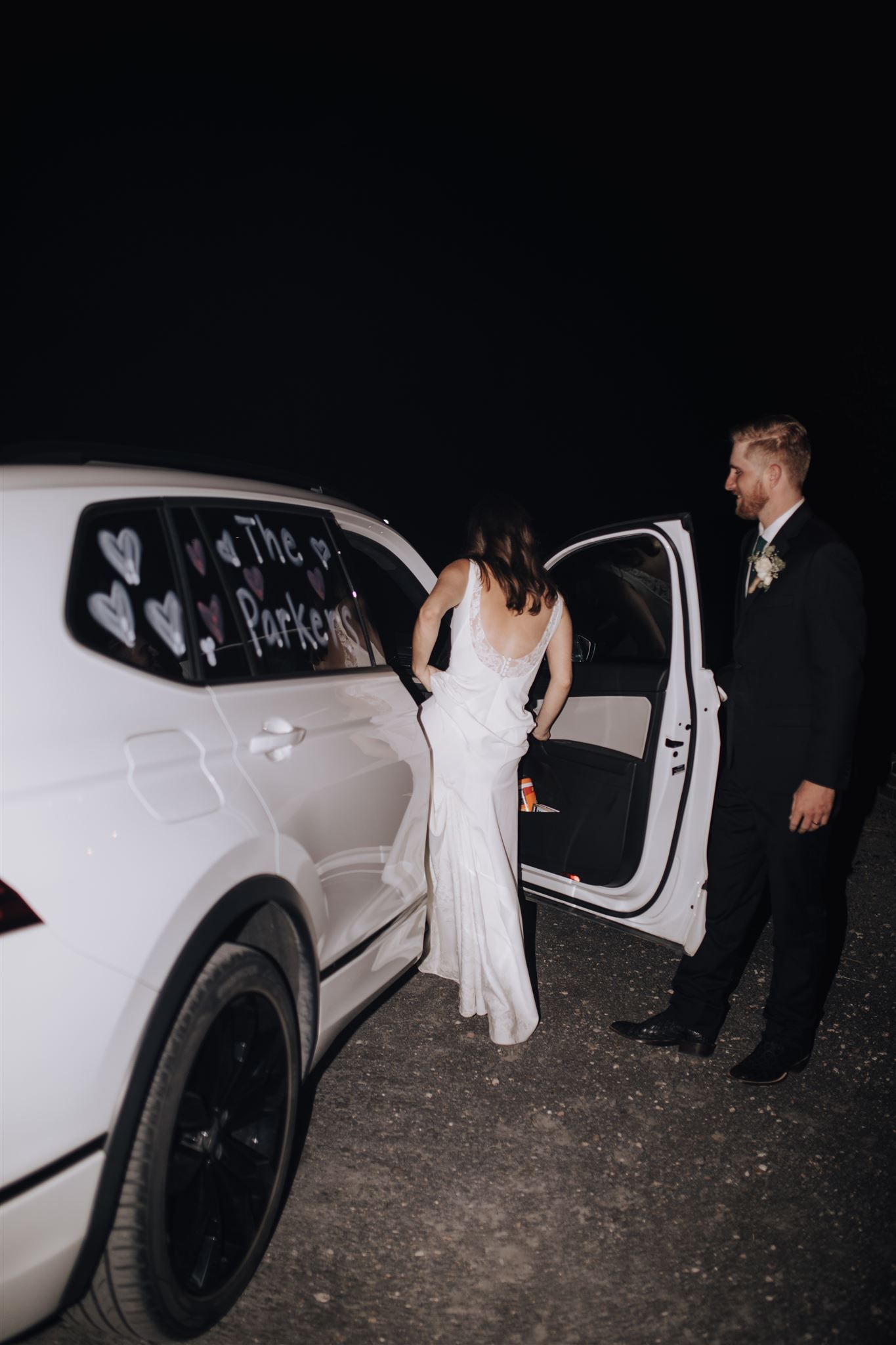 Couple departing wedding in white decorated getaway car at night.
