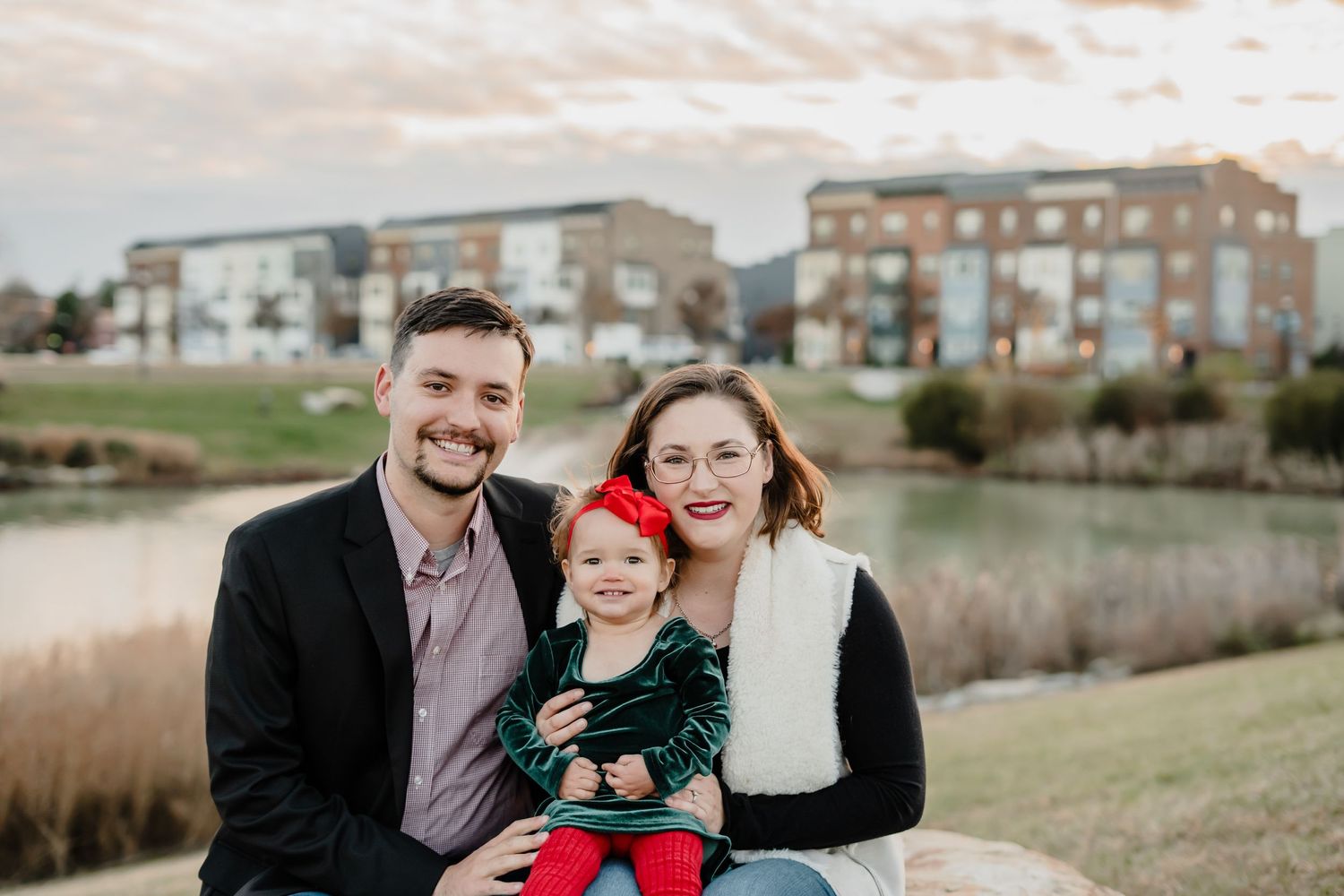 Family portrait by a pond during golden hour with urban buildings in background.