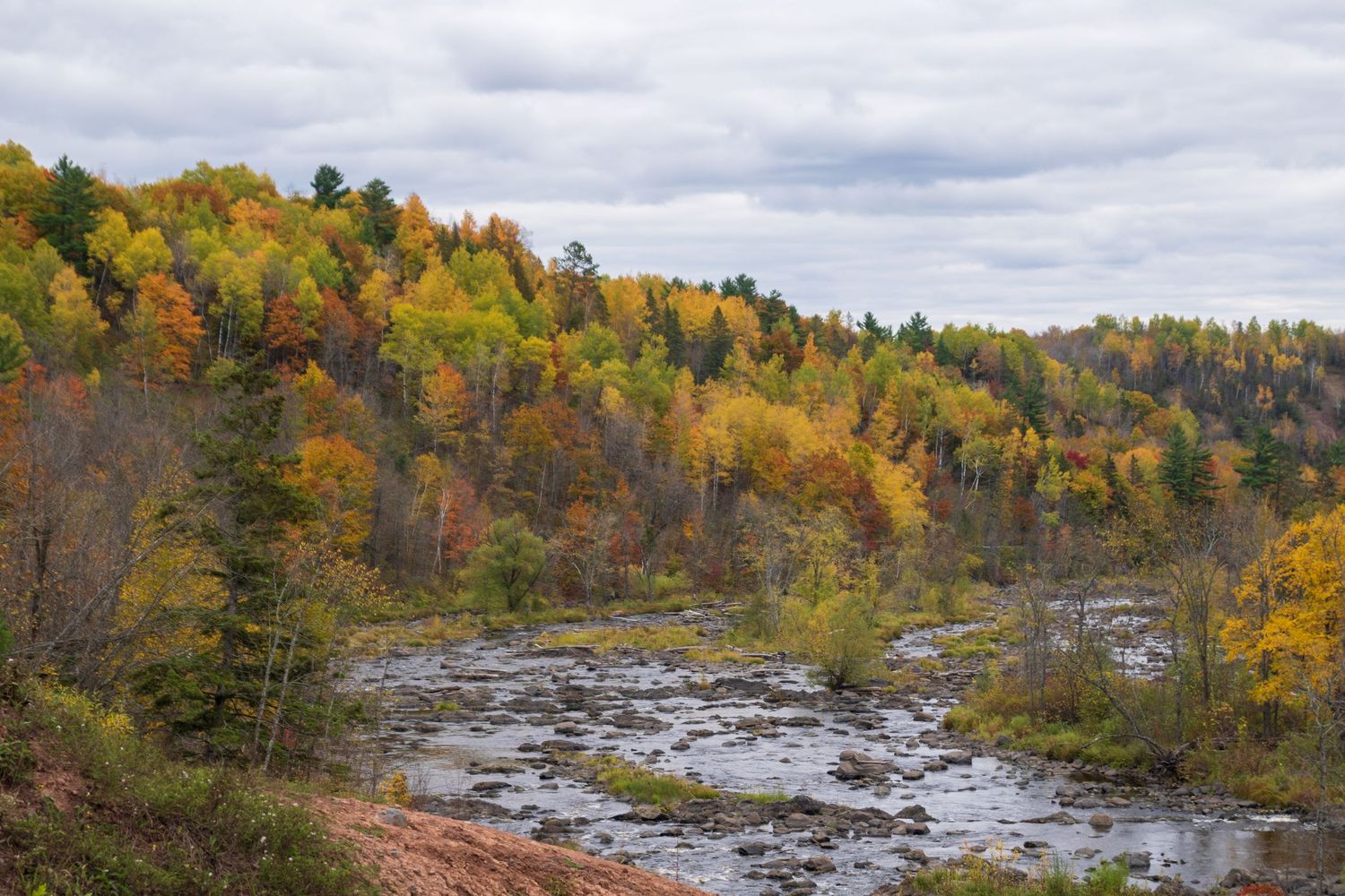 Autumn landscape of rocky riverbed surrounded by colorful fall foliage under cloudy sky.