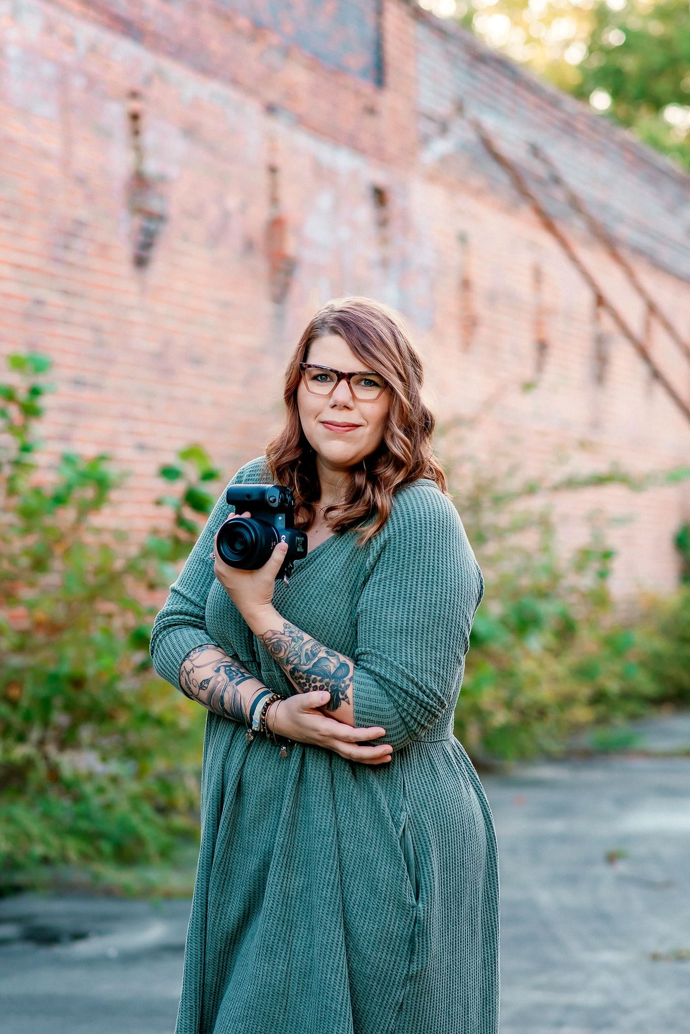 Professional photographer in a teal sweater holding a camera against a brick wall backdrop.