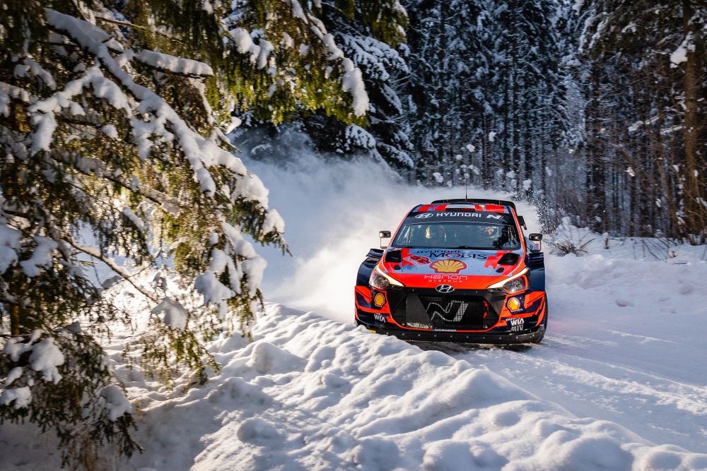Rally car races through snowy forest track kicking up powder snow.