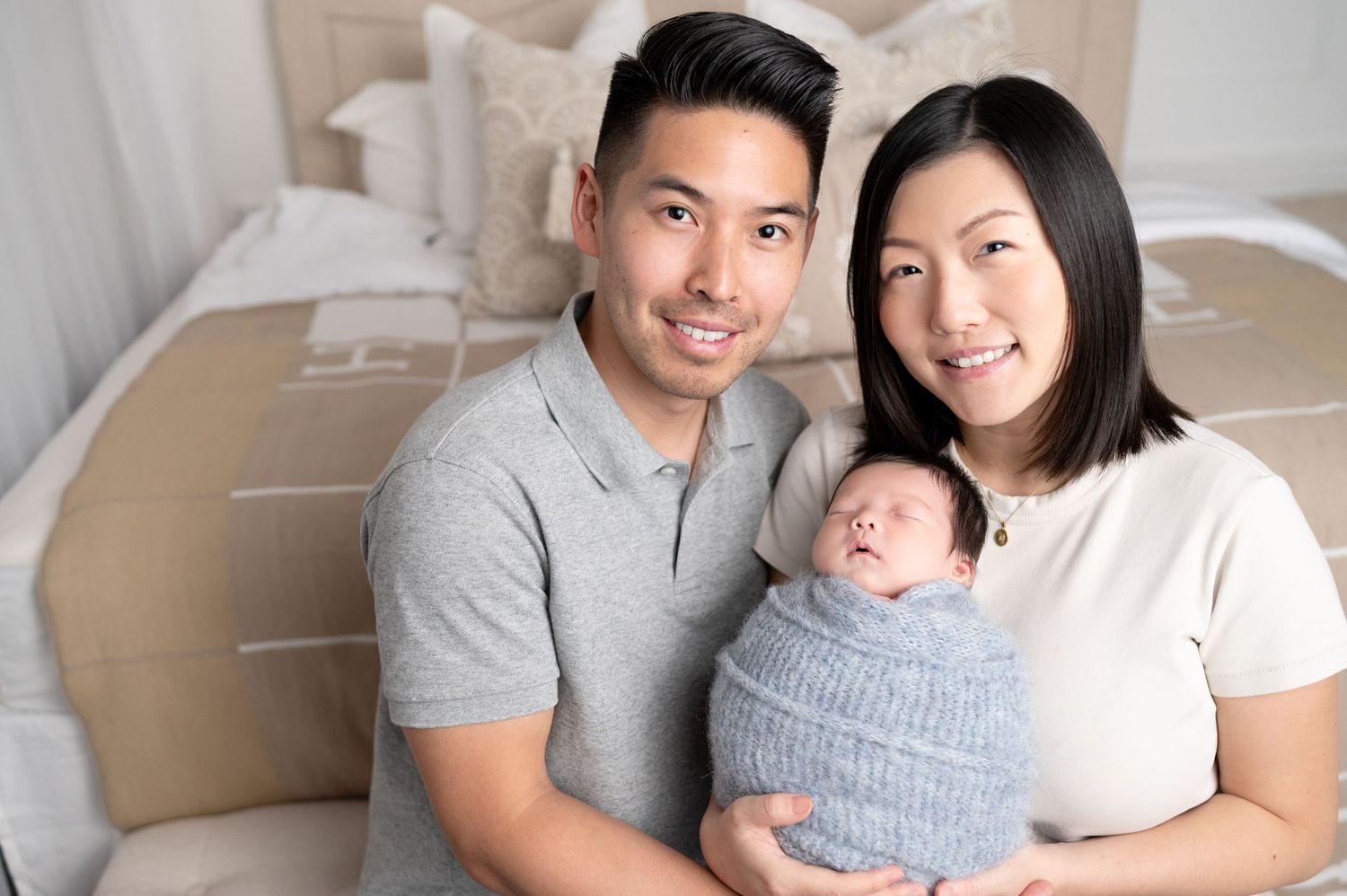 Family portrait with newborn baby wrapped in grey swaddle held by parents in casual attire.