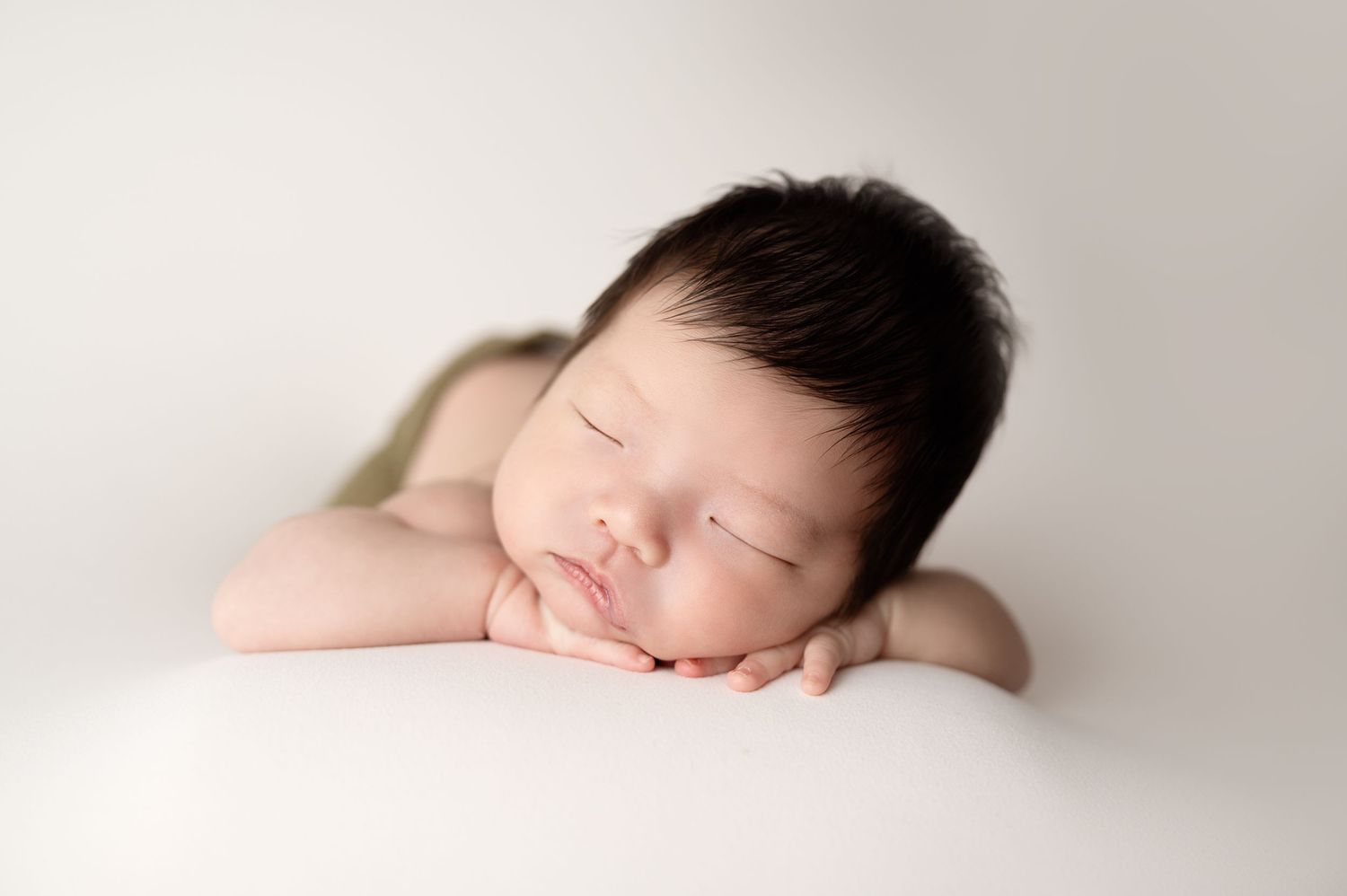 Newborn sleeping sweetly with head resting on crossed arms against white backdrop.