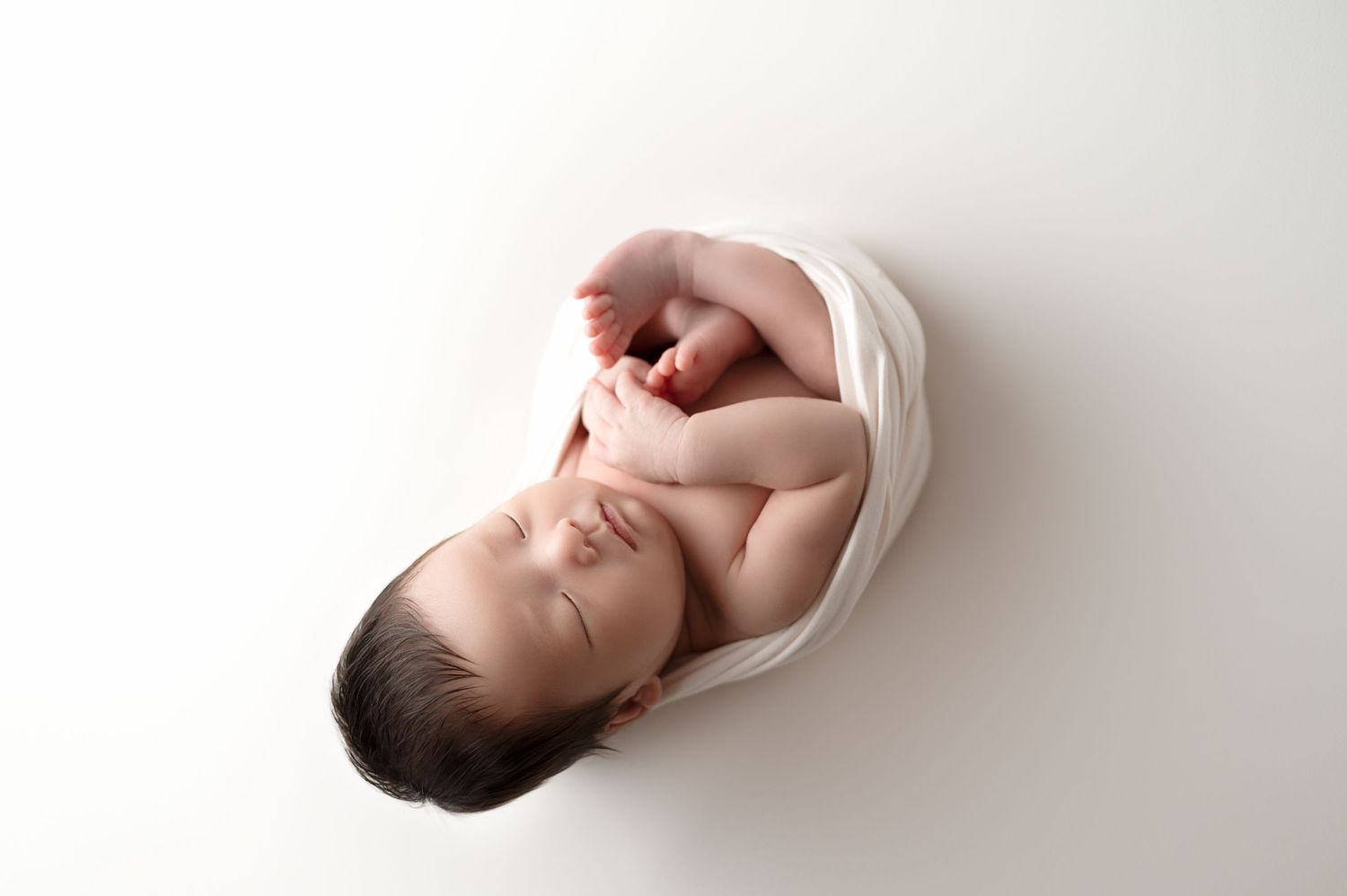 Sleeping newborn wrapped in white blanket on minimal white background.