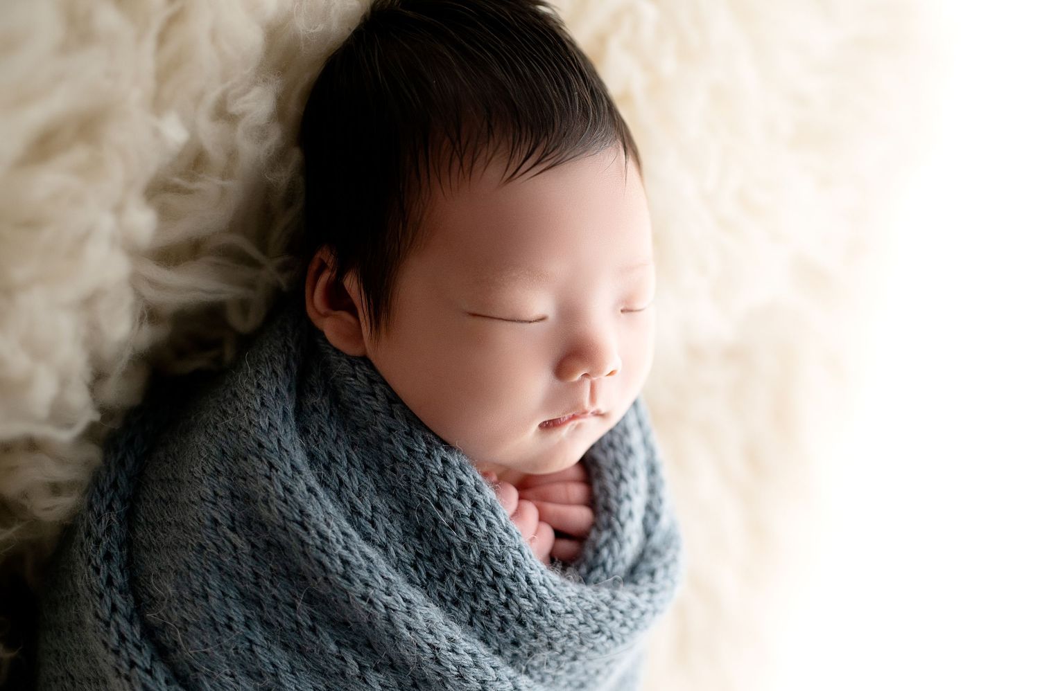 Baby sleeping peacefully wrapped in soft blue knitted blanket against cream background.