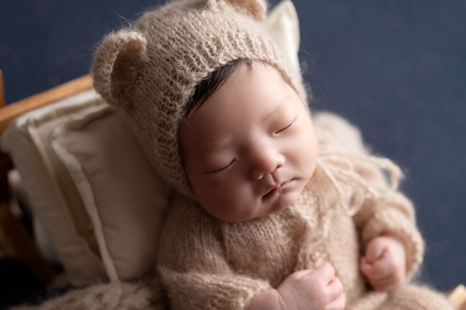 Sleeping newborn wearing cozy beige bear hat and knit outfit against dark backdrop.