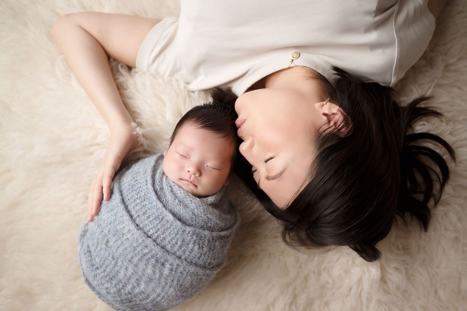 Mother and baby lying on floor, both peaceful with baby wrapped in grey knit blanket.