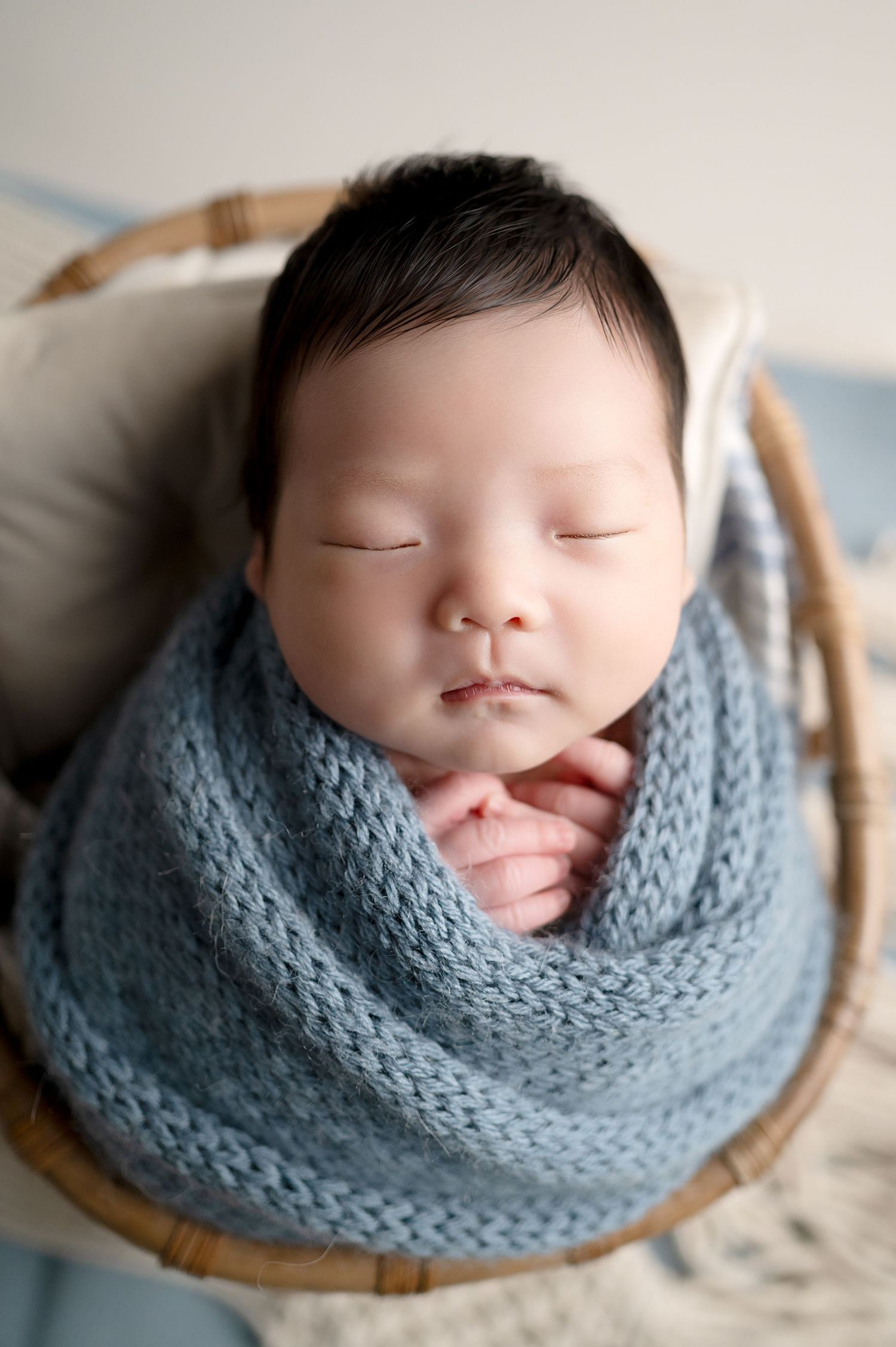 Newborn wrapped in blue knit blanket sleeping in wooden basket.